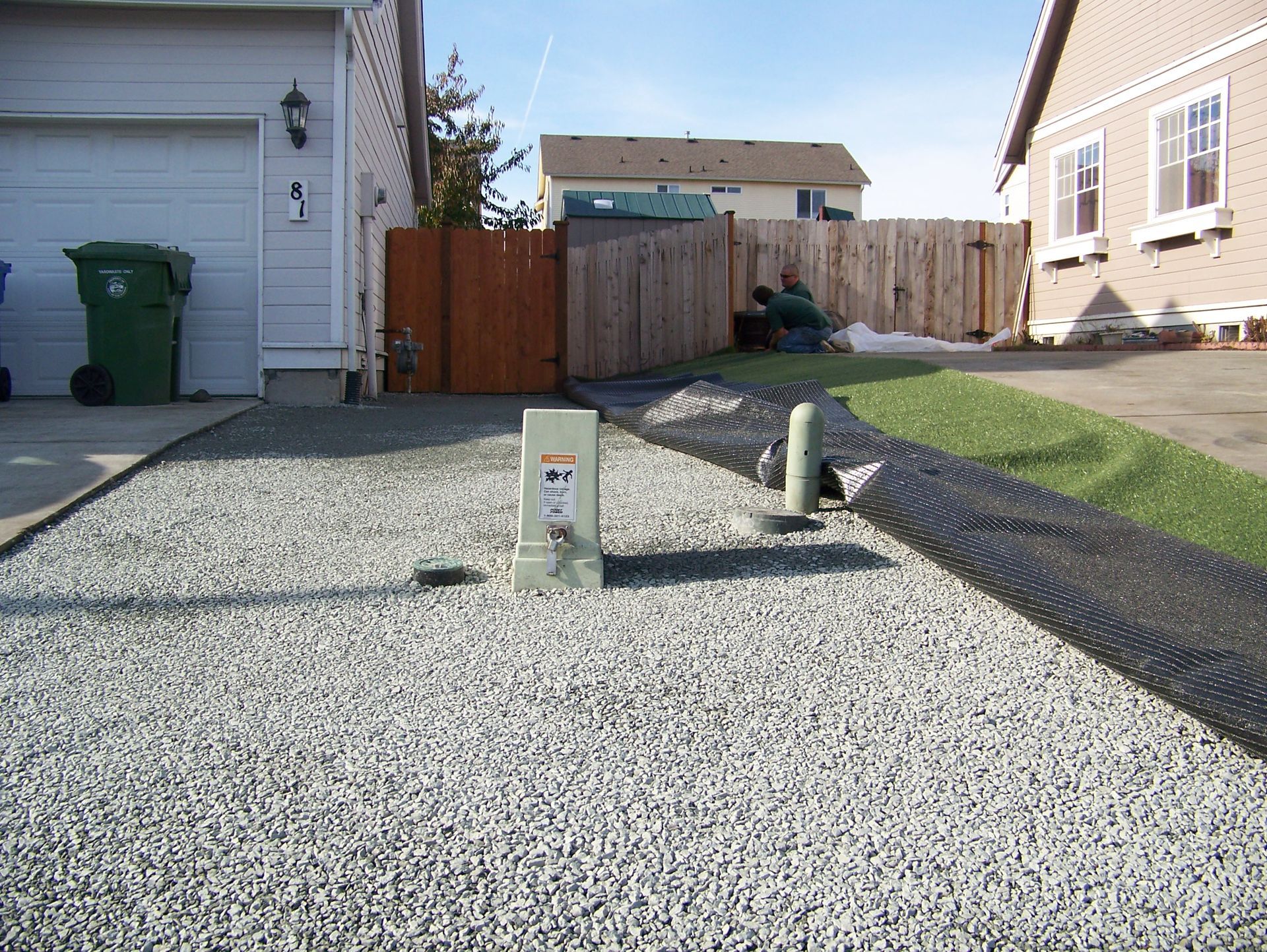 Gravel driveway and yard; power box, green trash can, and wooden fence; houses in the background.