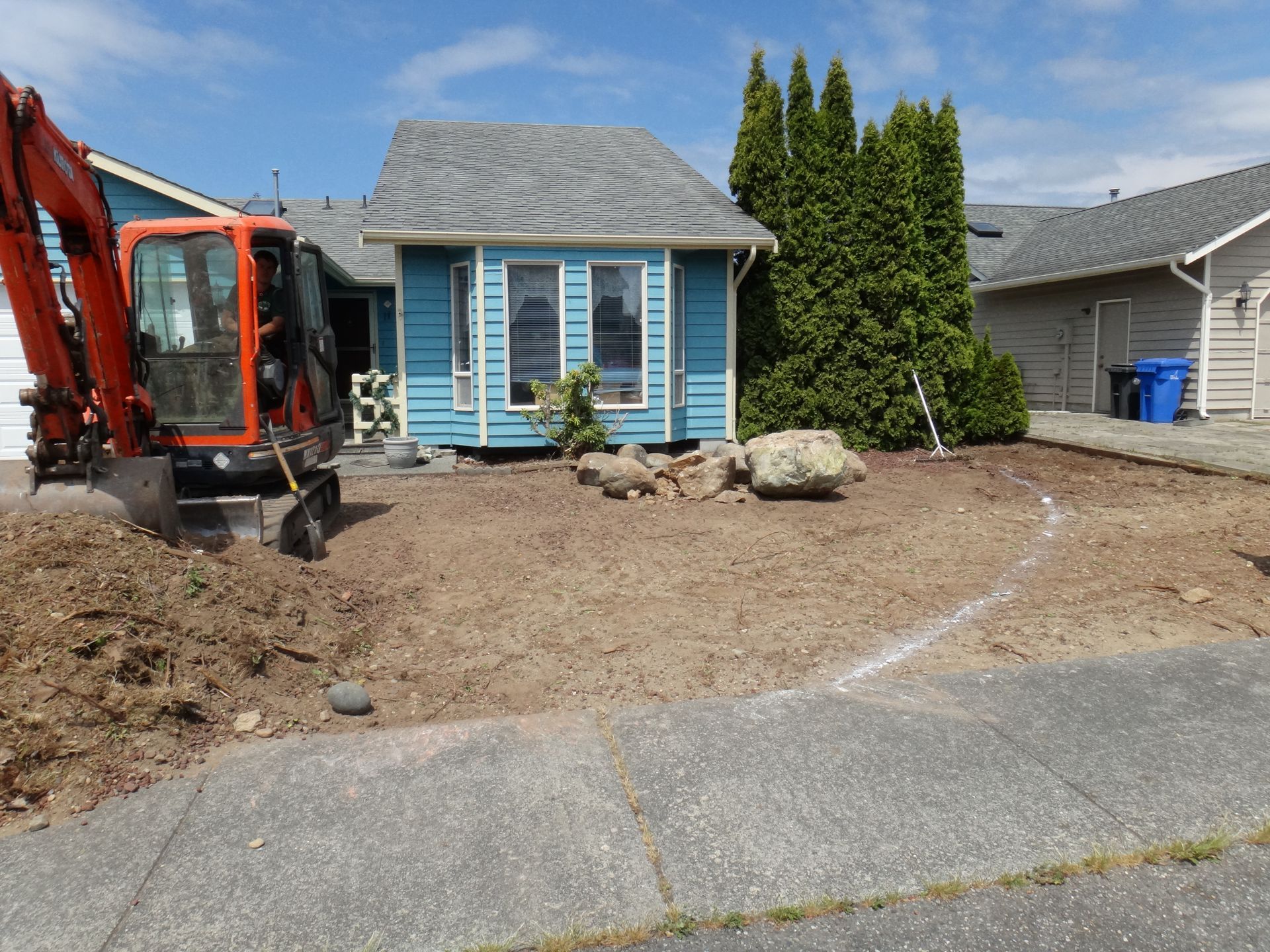 Mini excavator in front yard of blue house during landscaping.