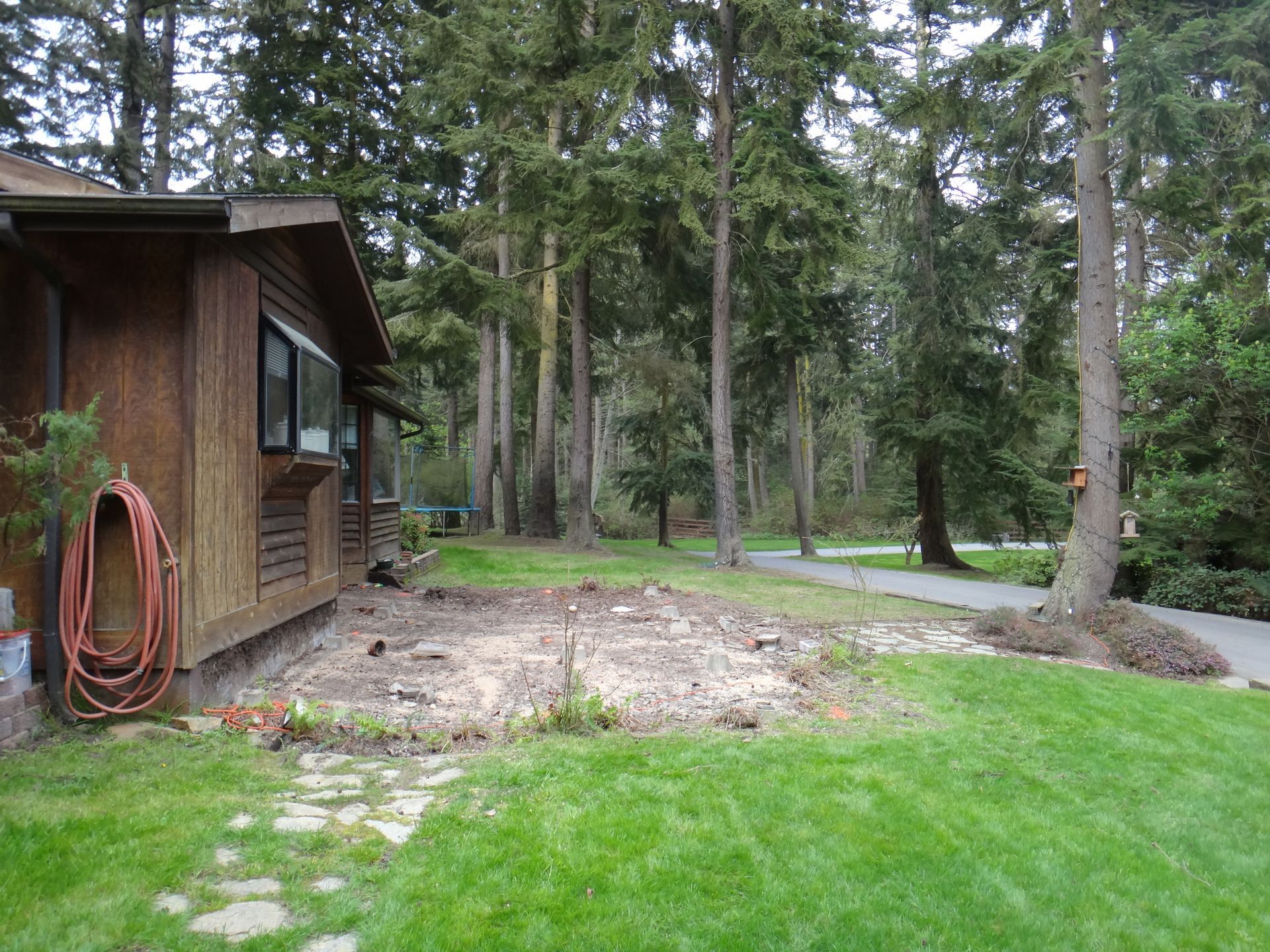 Side view of a brown house with a hose, next to a clearing surrounded by tall trees and a green lawn.