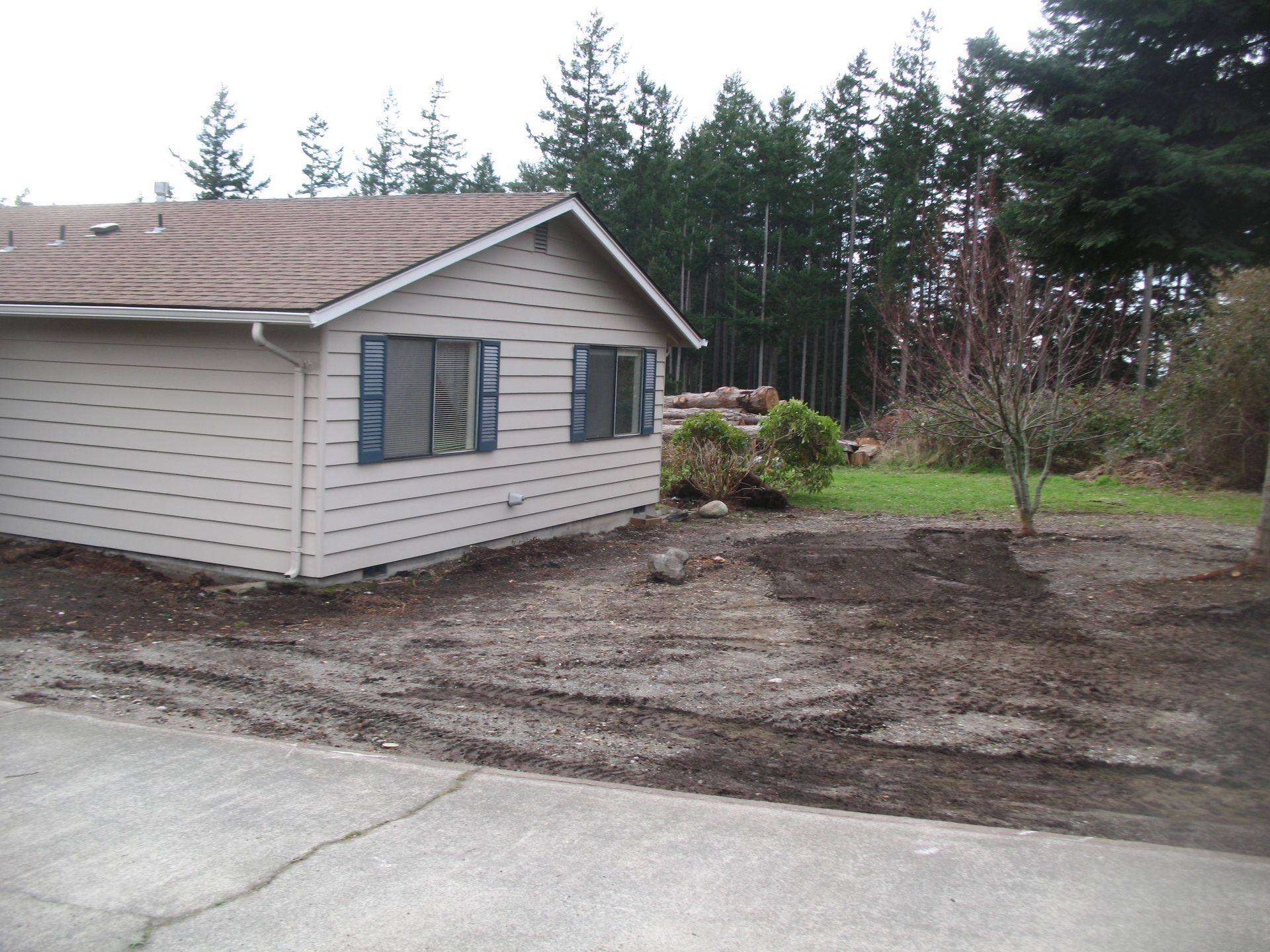 Beige house with blue shutters, dirt and tire tracks in front. Green grass and trees in background.