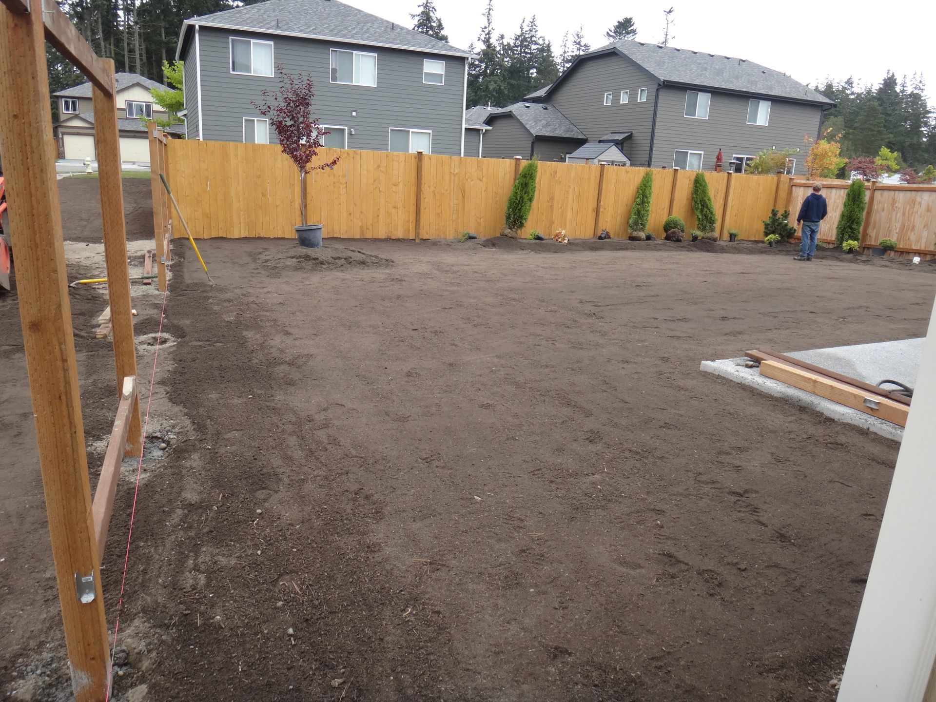 A backyard with freshly tilled soil, a wooden fence, and a person. Gray houses in the background.
