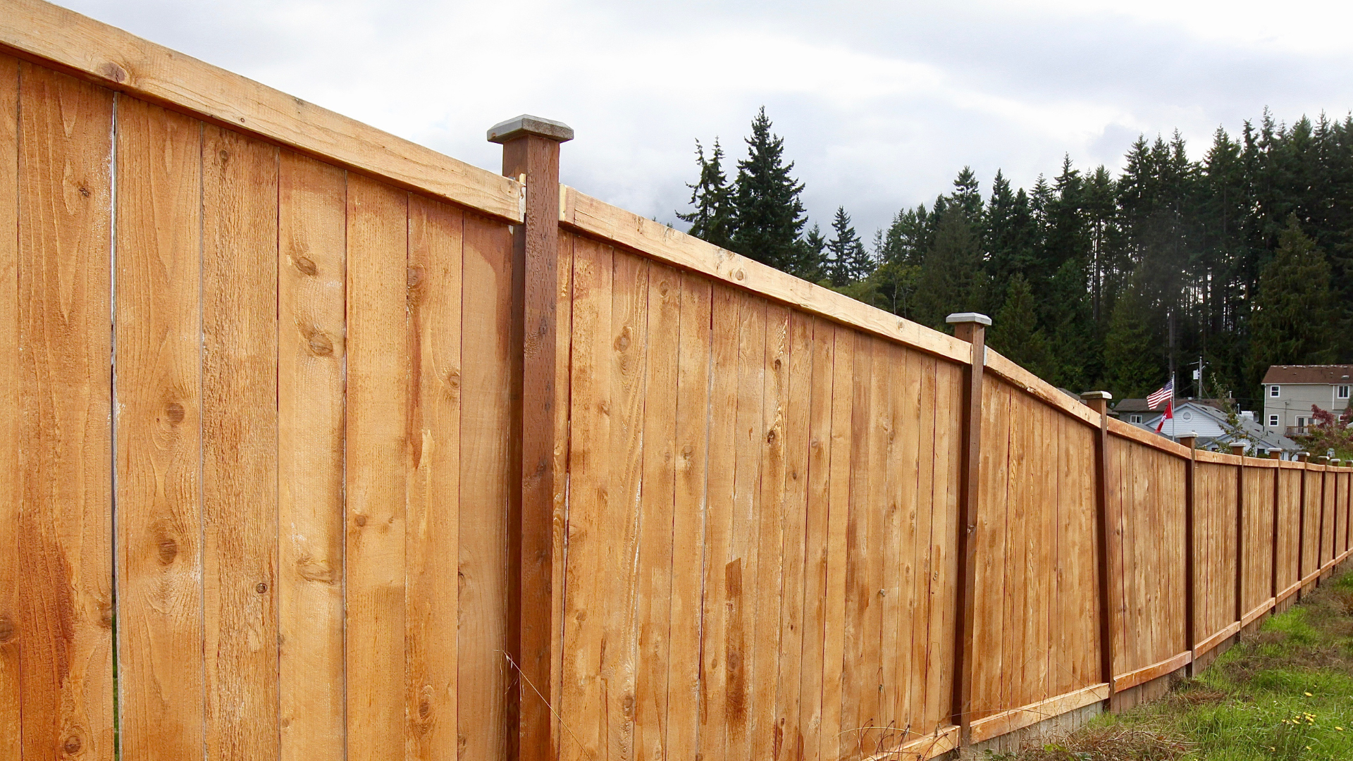 Wooden fence extending along a grassy area, with trees and a cloudy sky in the background.