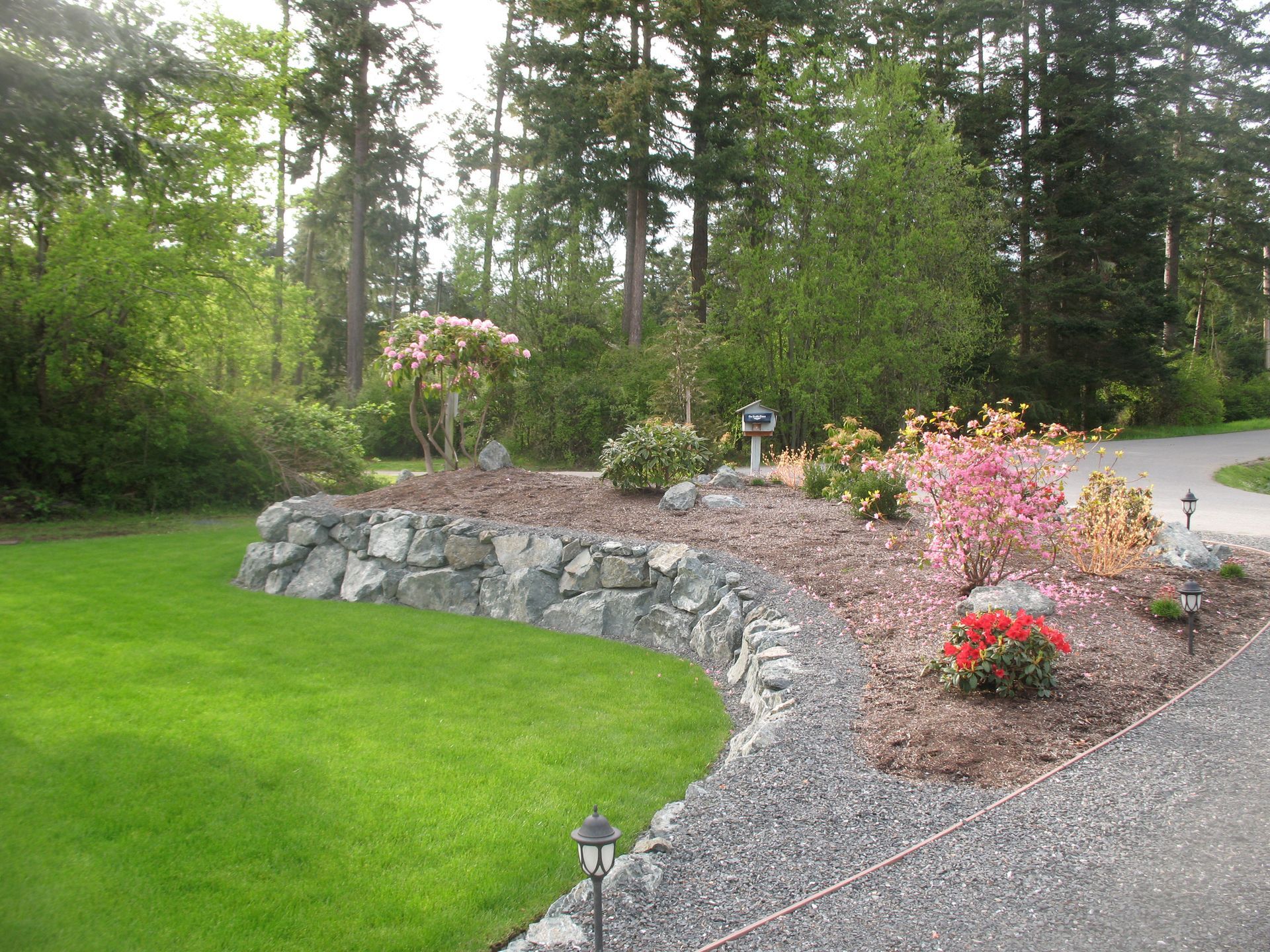 Curving stone wall in garden with green lawn, gravel path, and colorful blooming plants.