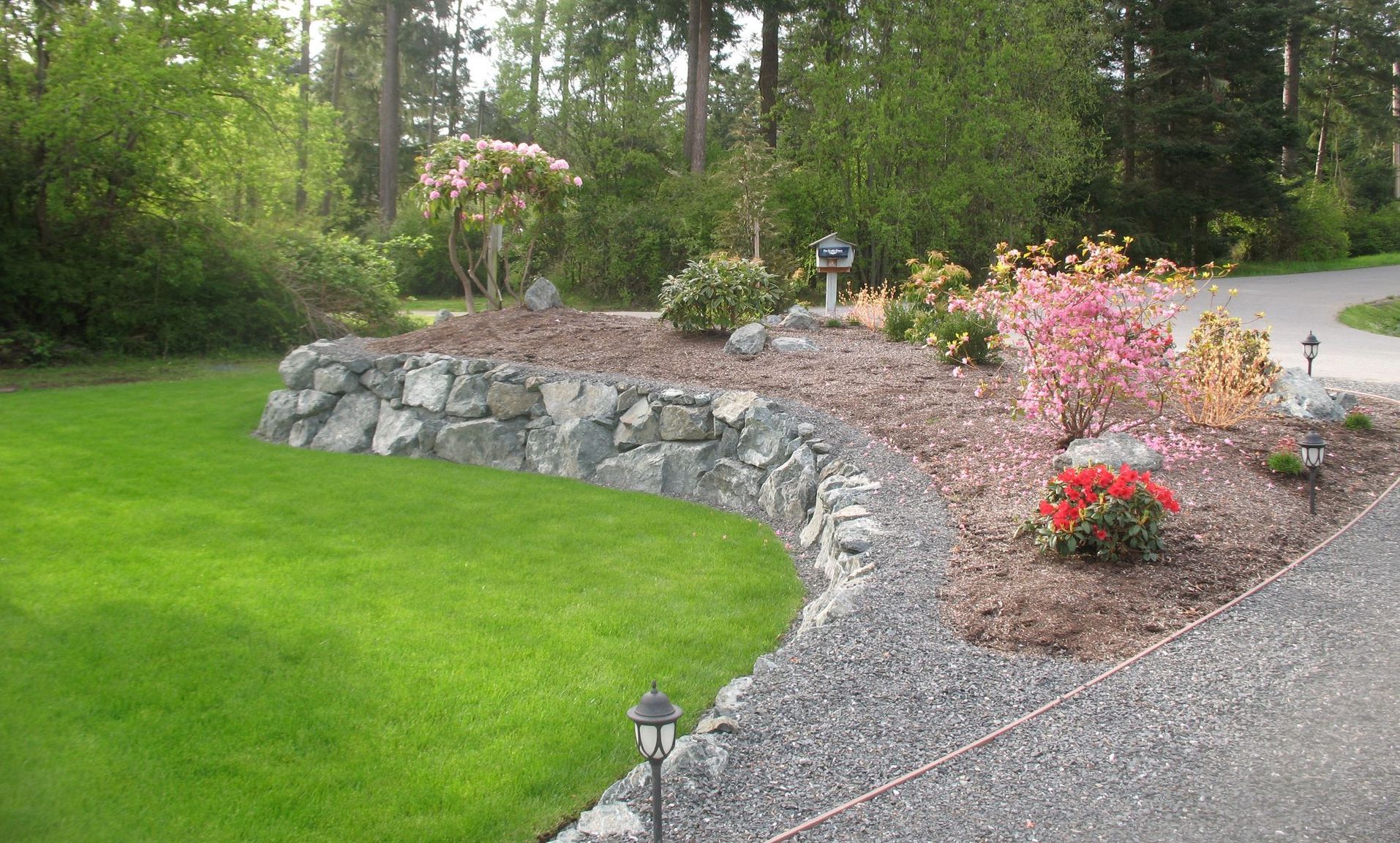 Curved stone retaining wall with garden beds of colorful plants next to a driveway and green lawn.