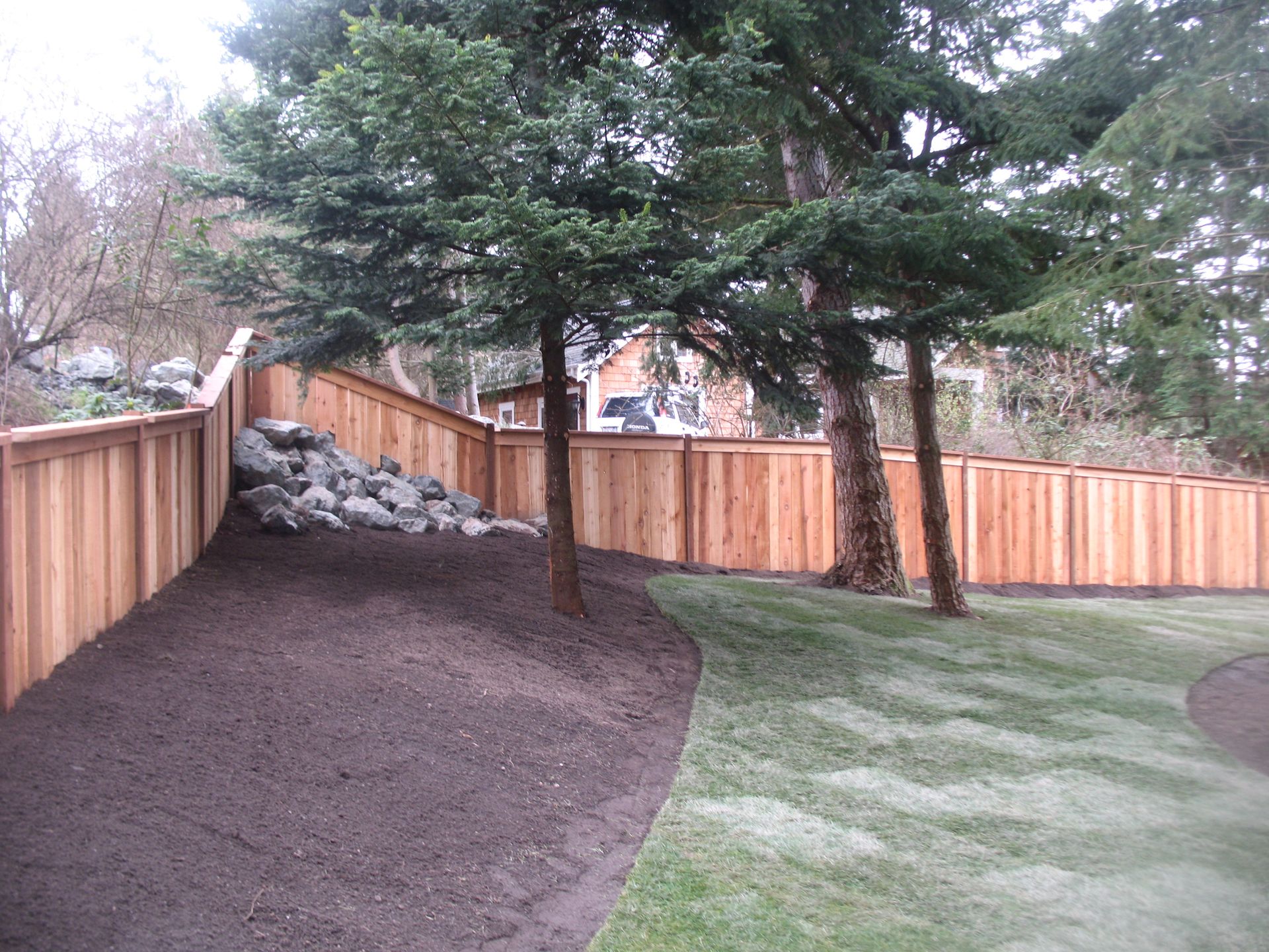 Wooden fence along a slight hillside with trees and grass, newly mulched area.