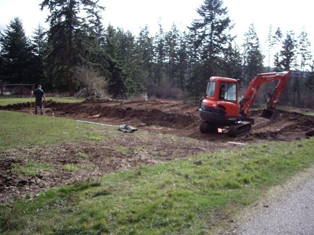 Mini excavator digging in a dirt area, person standing in the background.