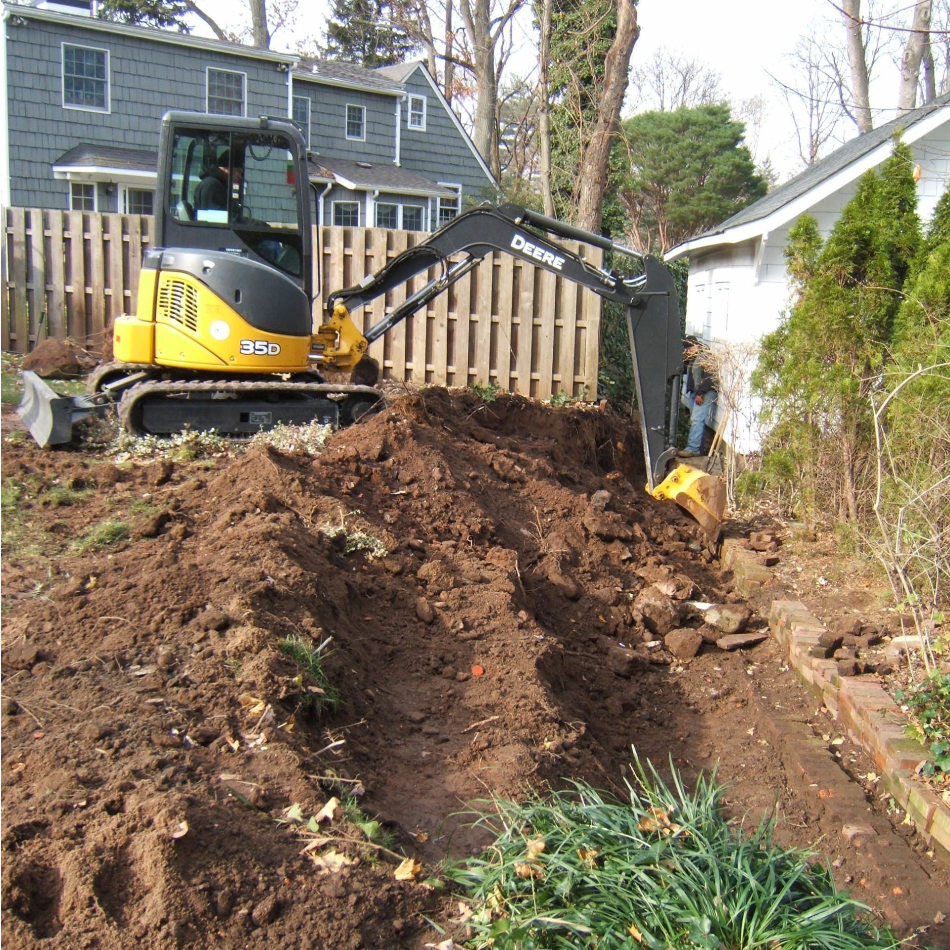 Yellow excavator digging in a residential backyard; piles of dirt, wooden fence, house in background.