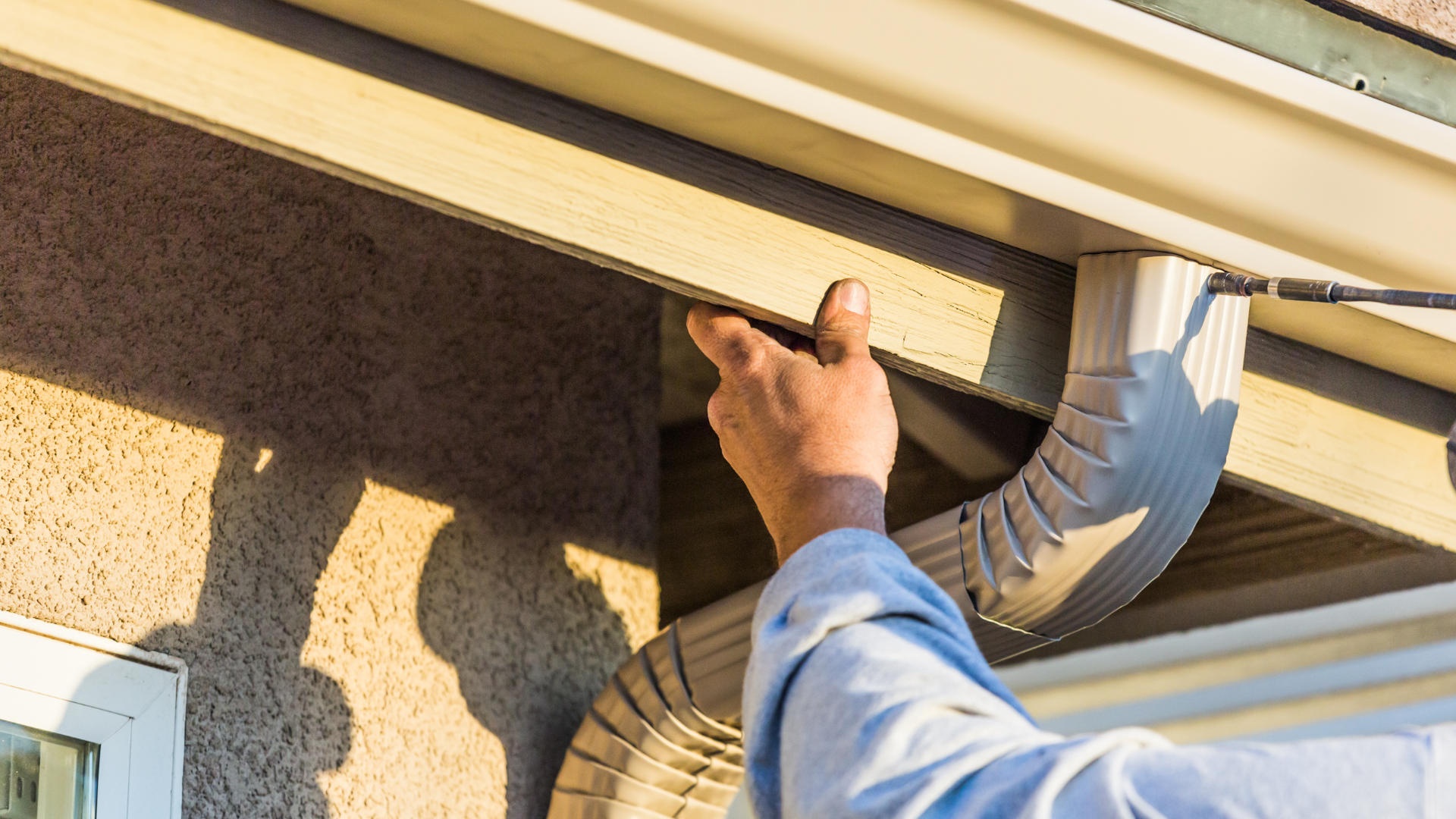 Person installing a light beige gutter on a building's exterior, using a tool.