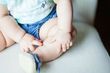 Baby in jean shorts and blue shoes sits on a white chair, touching legs.