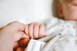Baby's hand holding an adult's finger; both are flesh-toned, close-up, indoors.