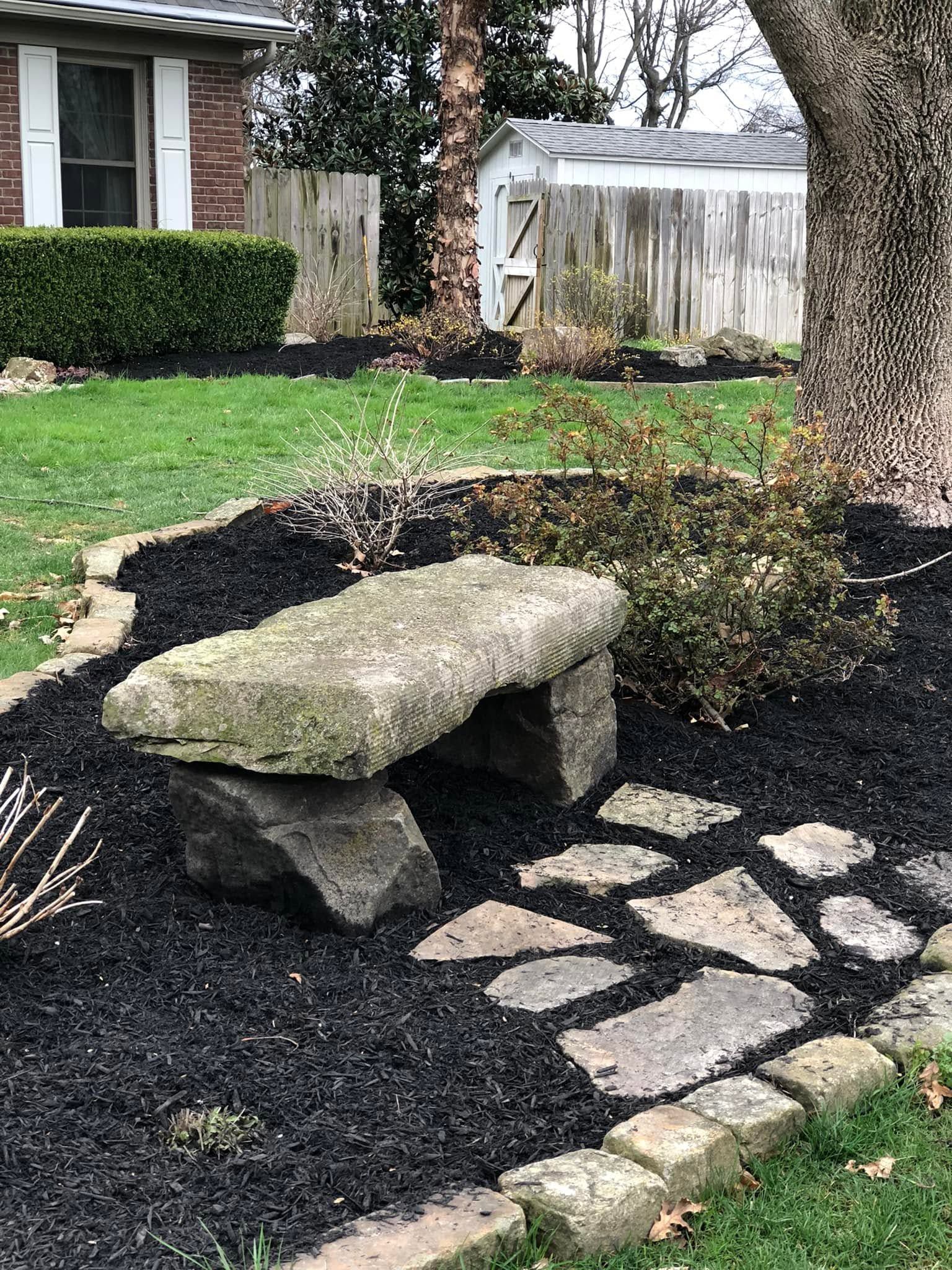 Garden path with stone bench, black mulch, and stepping stones near a tree in a suburban yard