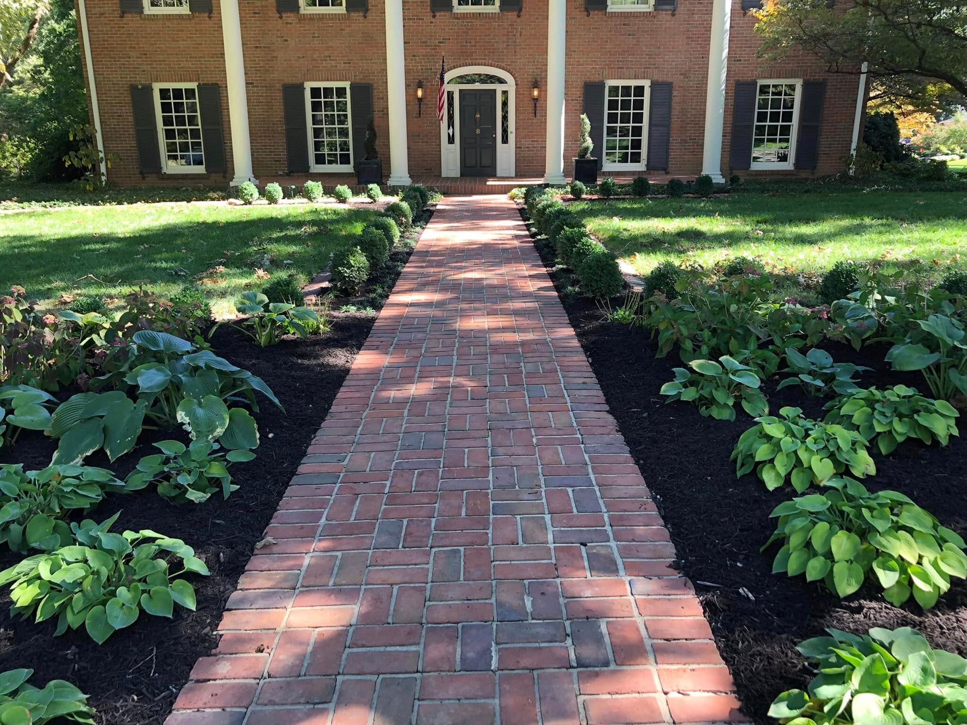 Brick walkway leading to a brick house entrance, bordered by green shrubs and lawn.