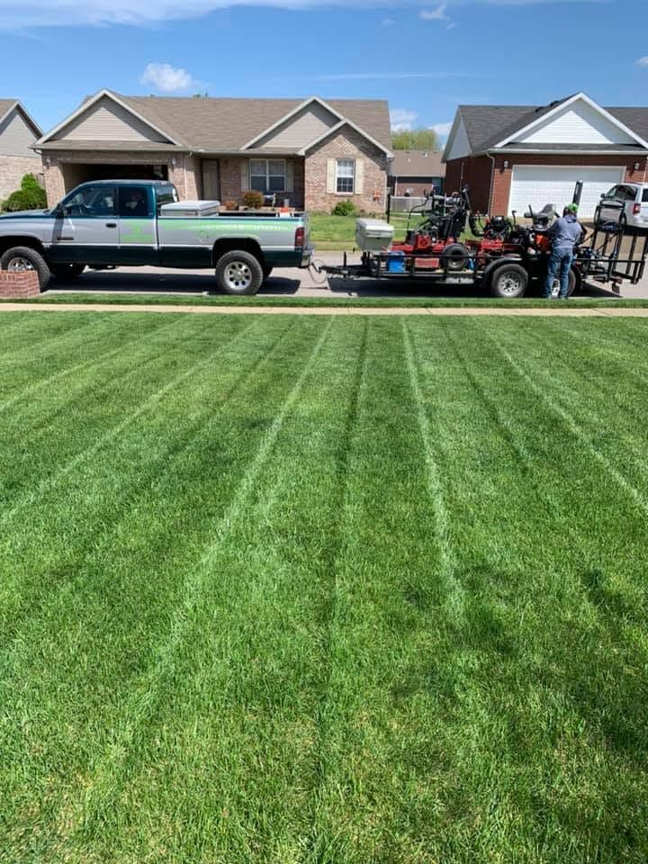 Suburban street with a green pickup, lawn mower trailer, and workers on a striped green lawn under a blue sky