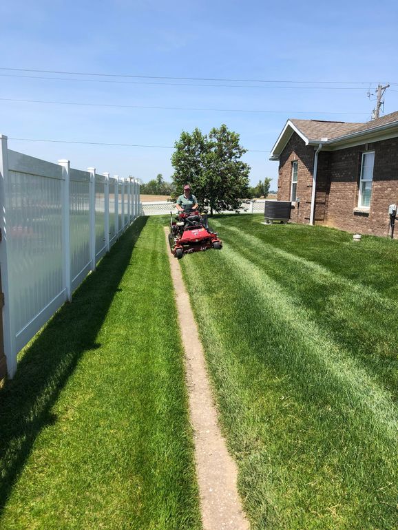Red lawn tractor on striped grass beside a white fence and brick house under a clear blue sky