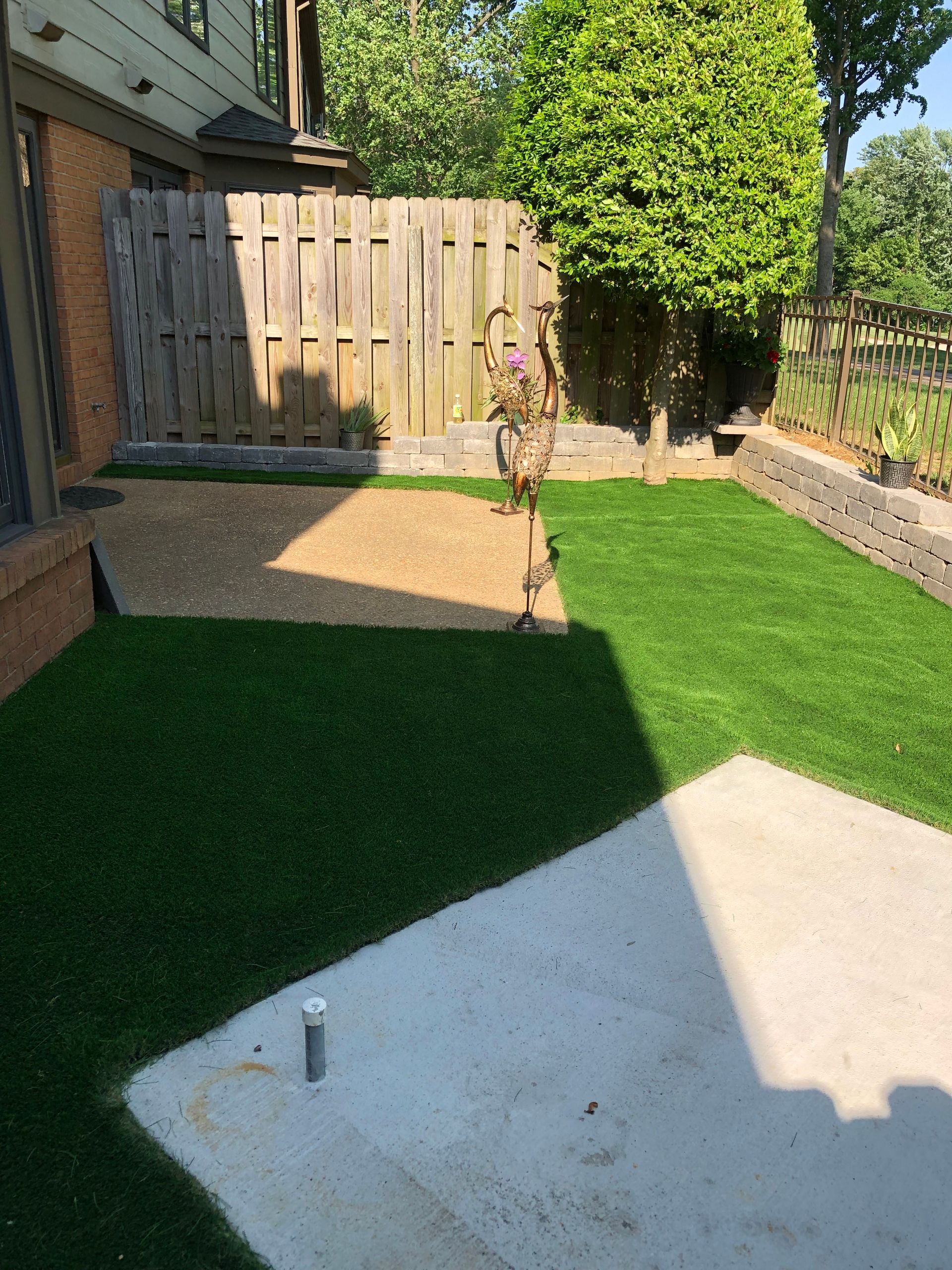 Backyard with artificial turf, patio pavers, wooden fence, and a young tree in the center.