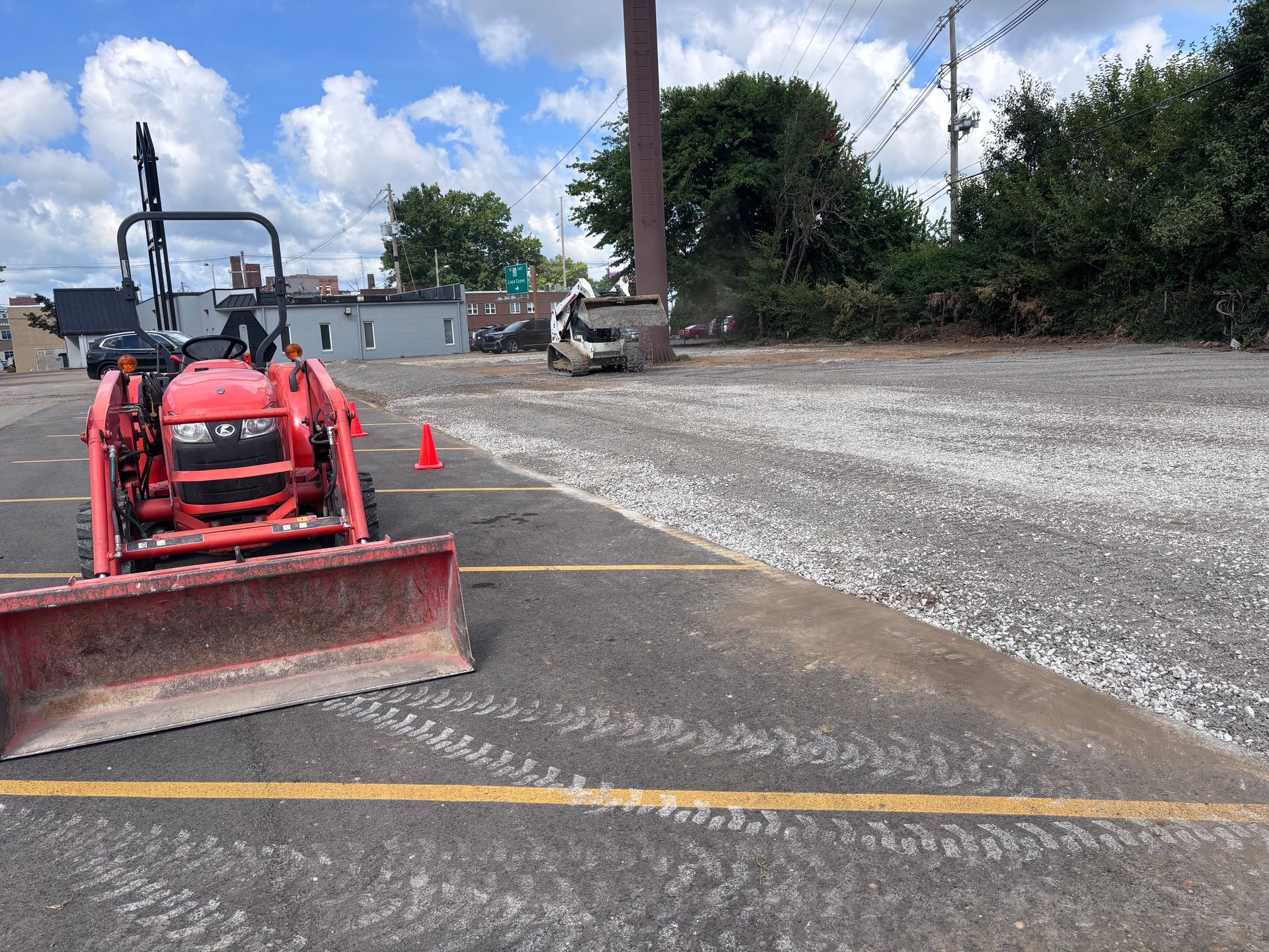 Red tractor with front blade on a gravel lot near cones, trees, and parked vehicles under a blue sky.