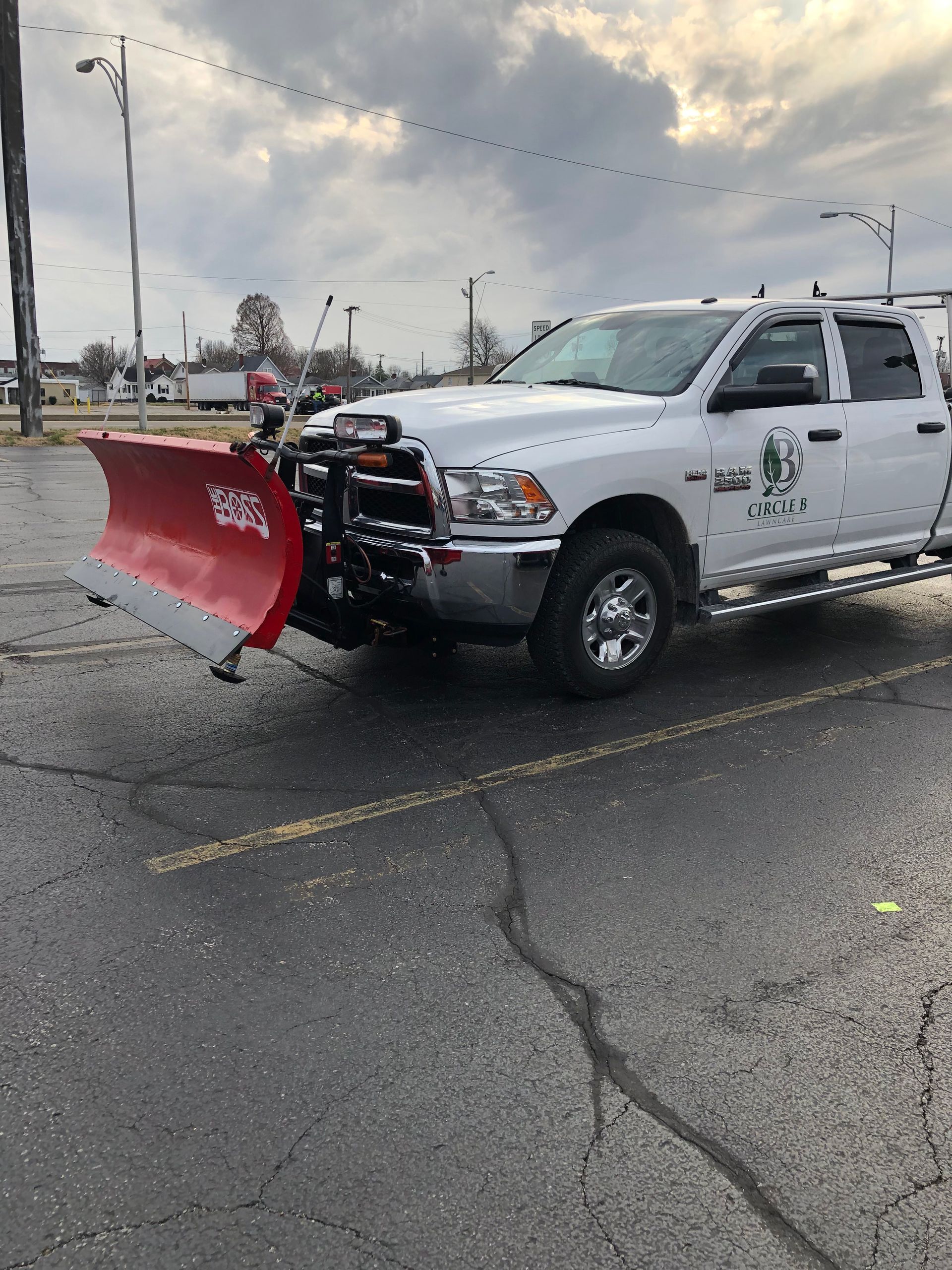 White pickup truck with a red snowplow attached, parked on a wet street under an overcast sky.