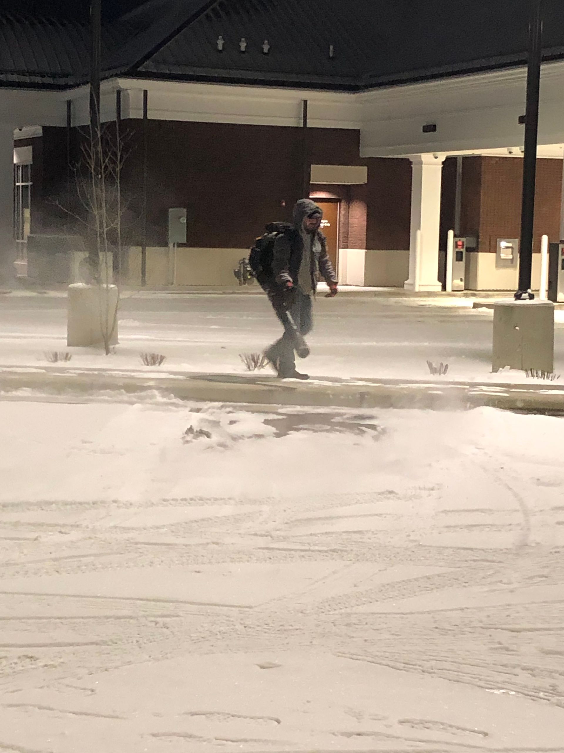 Two people walking through a snowy parking lot at night near a building, one carrying a backpack