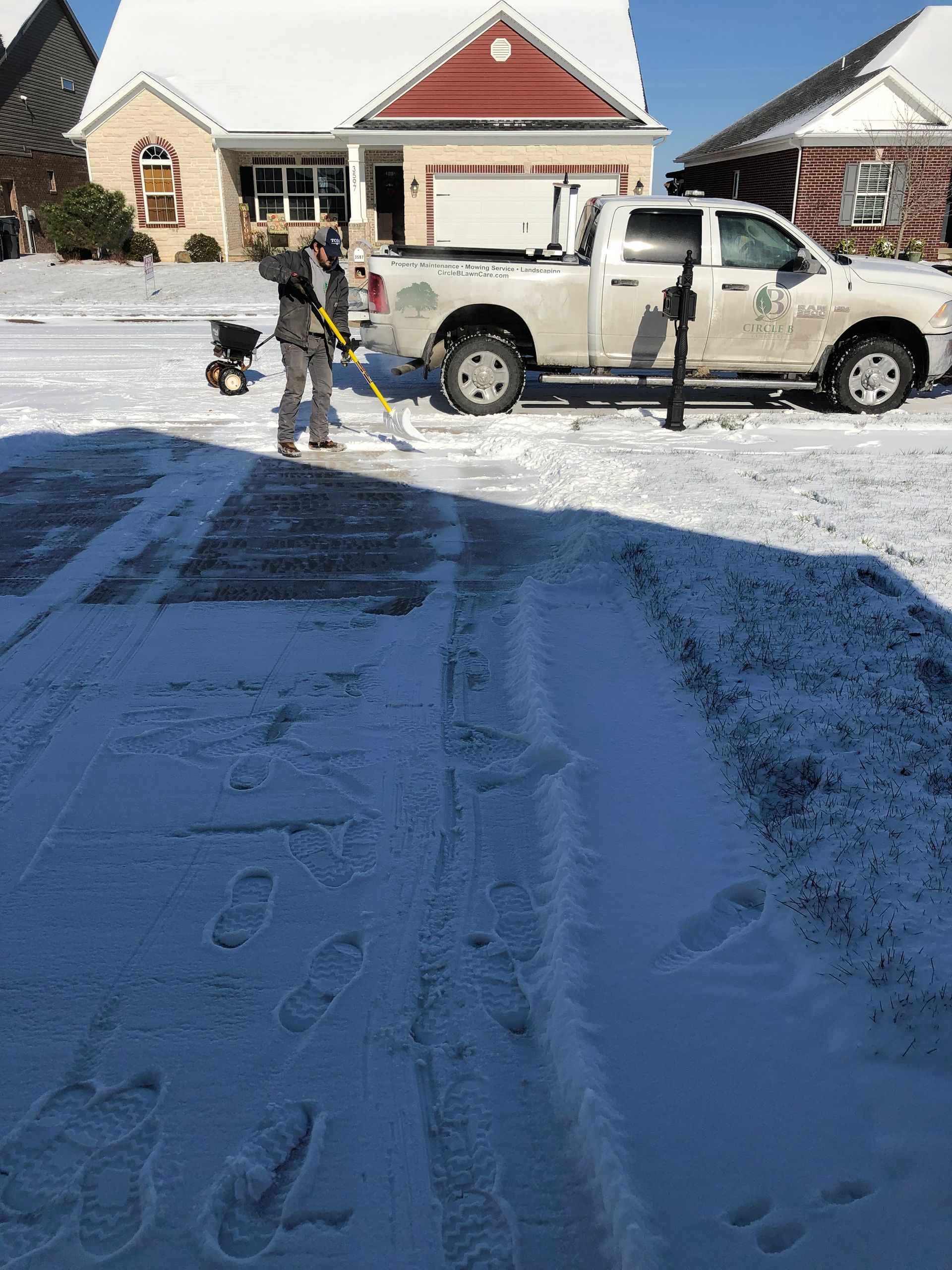 Person snow-blowing a driveway beside a white pickup truck in front of a house.