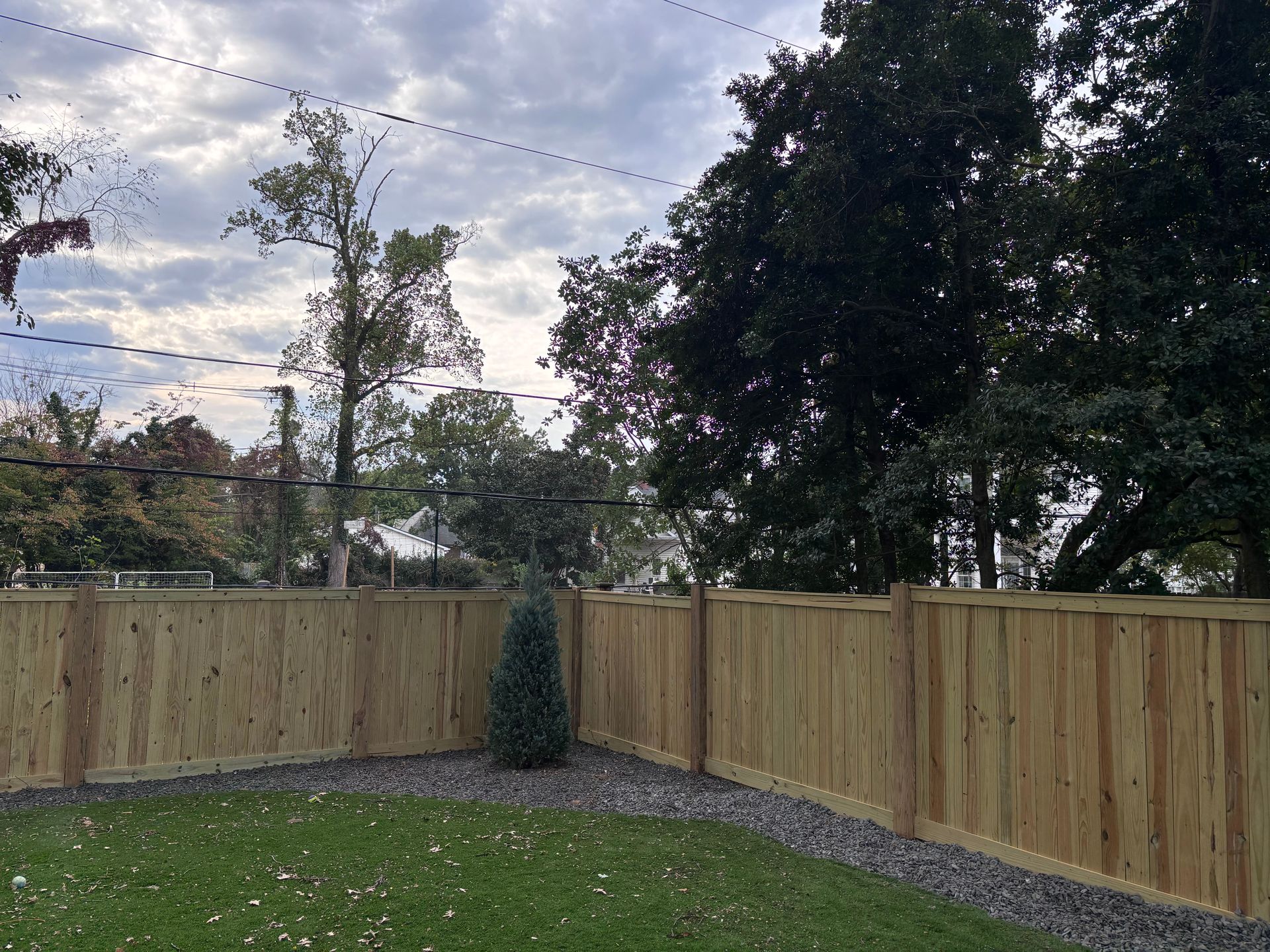 Backyard with wooden fence, green lawn, gravel border, small evergreen, and cloudy sky