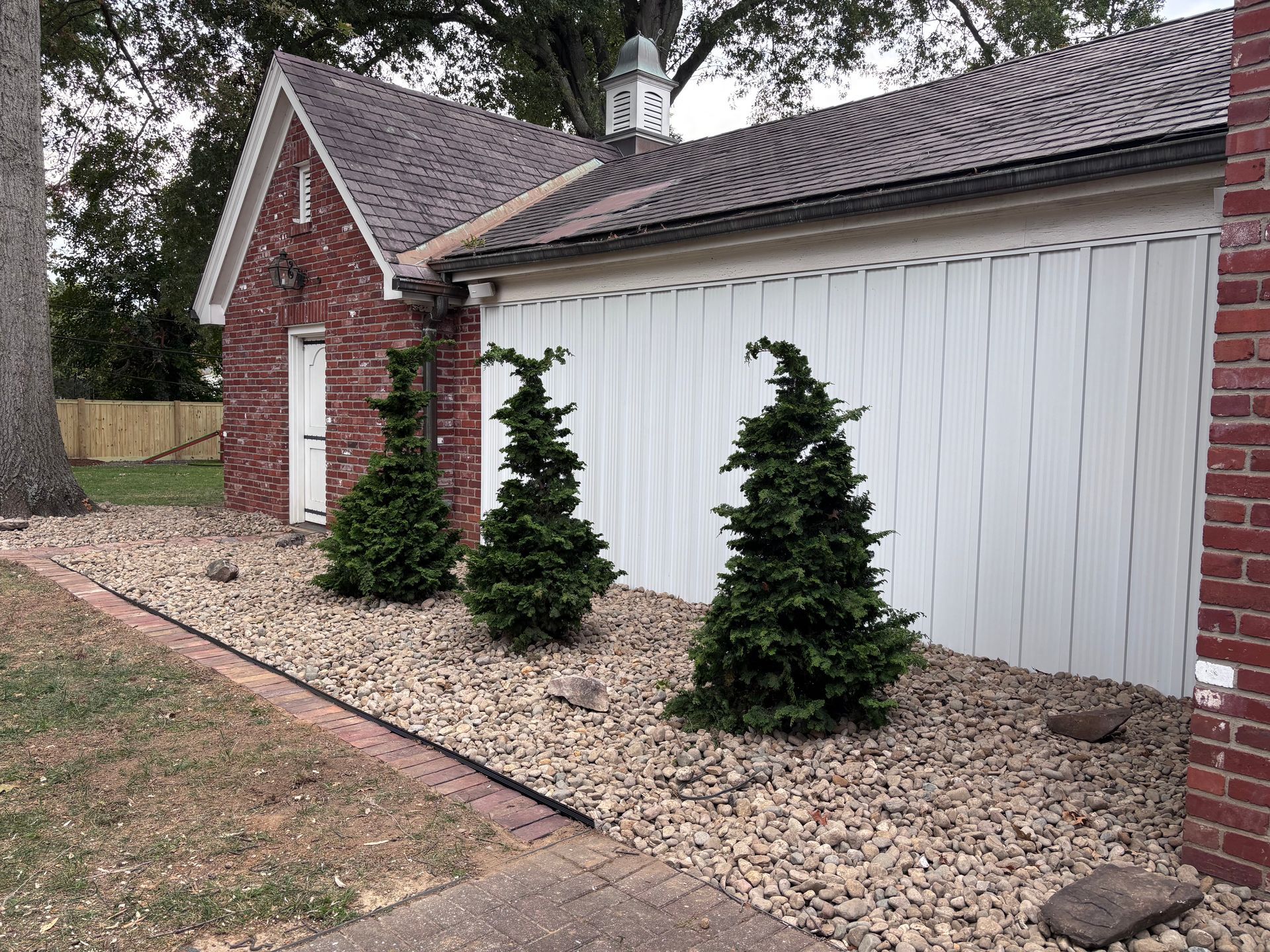 Brick garage with white doors, rock landscaping, and three small evergreen trees along the side