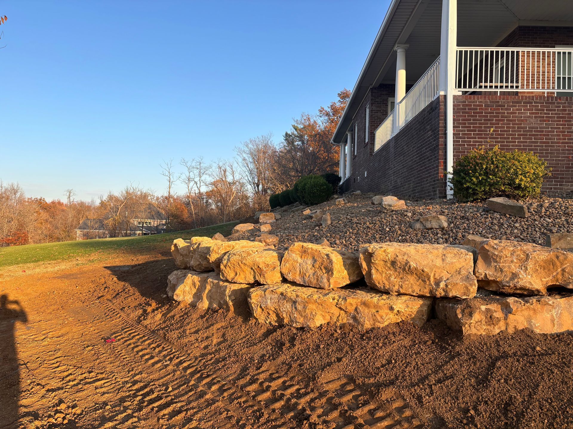 Stone retaining wall beside a brick house on a sunny hillside with blue sky and autumn trees