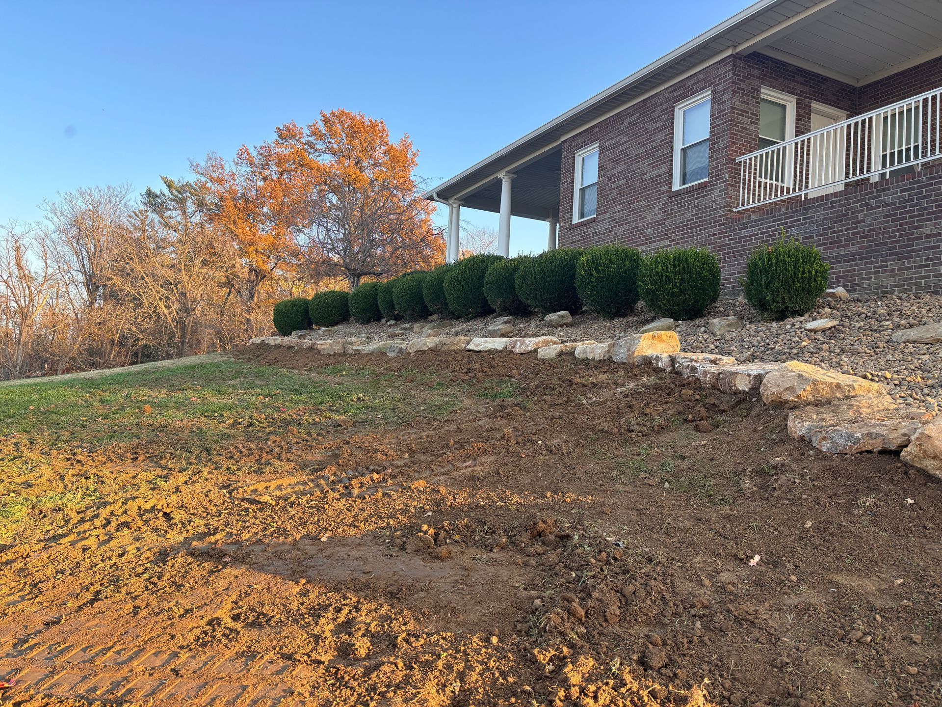 Brick house with trimmed shrubs on a sloped yard beside autumn trees at sunset