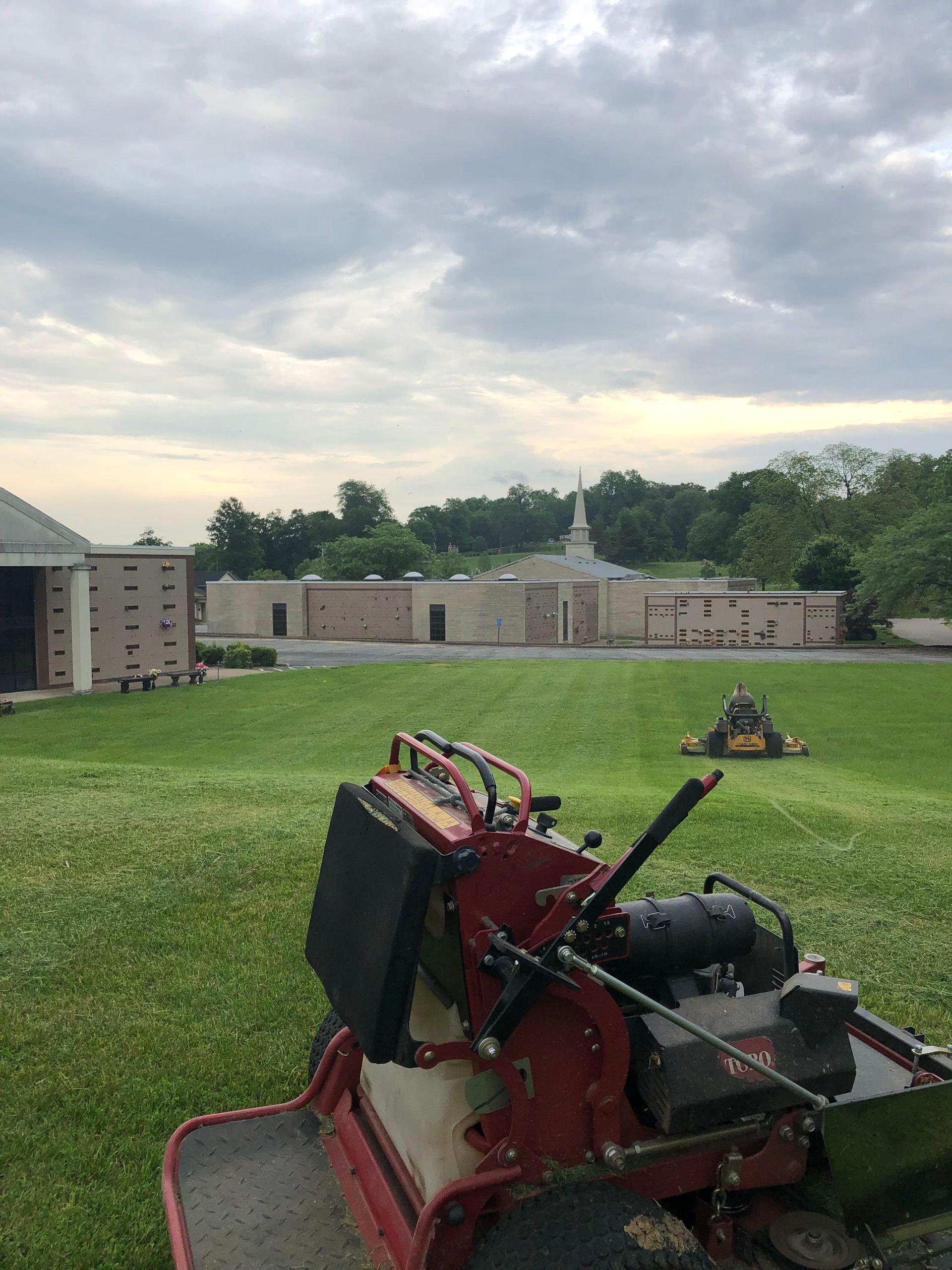 Red lawn mower on a grassy field with buildings and cloudy sky in the background