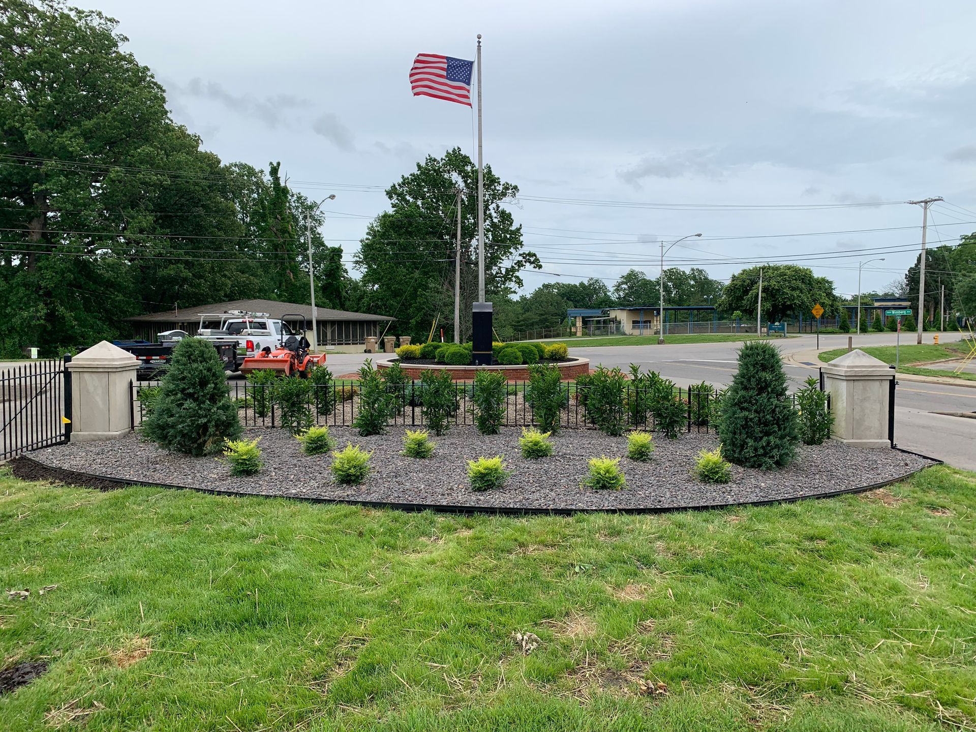 Landscaped memorial with small shrubs, an American flag, and a stone entrance gate by a roadside