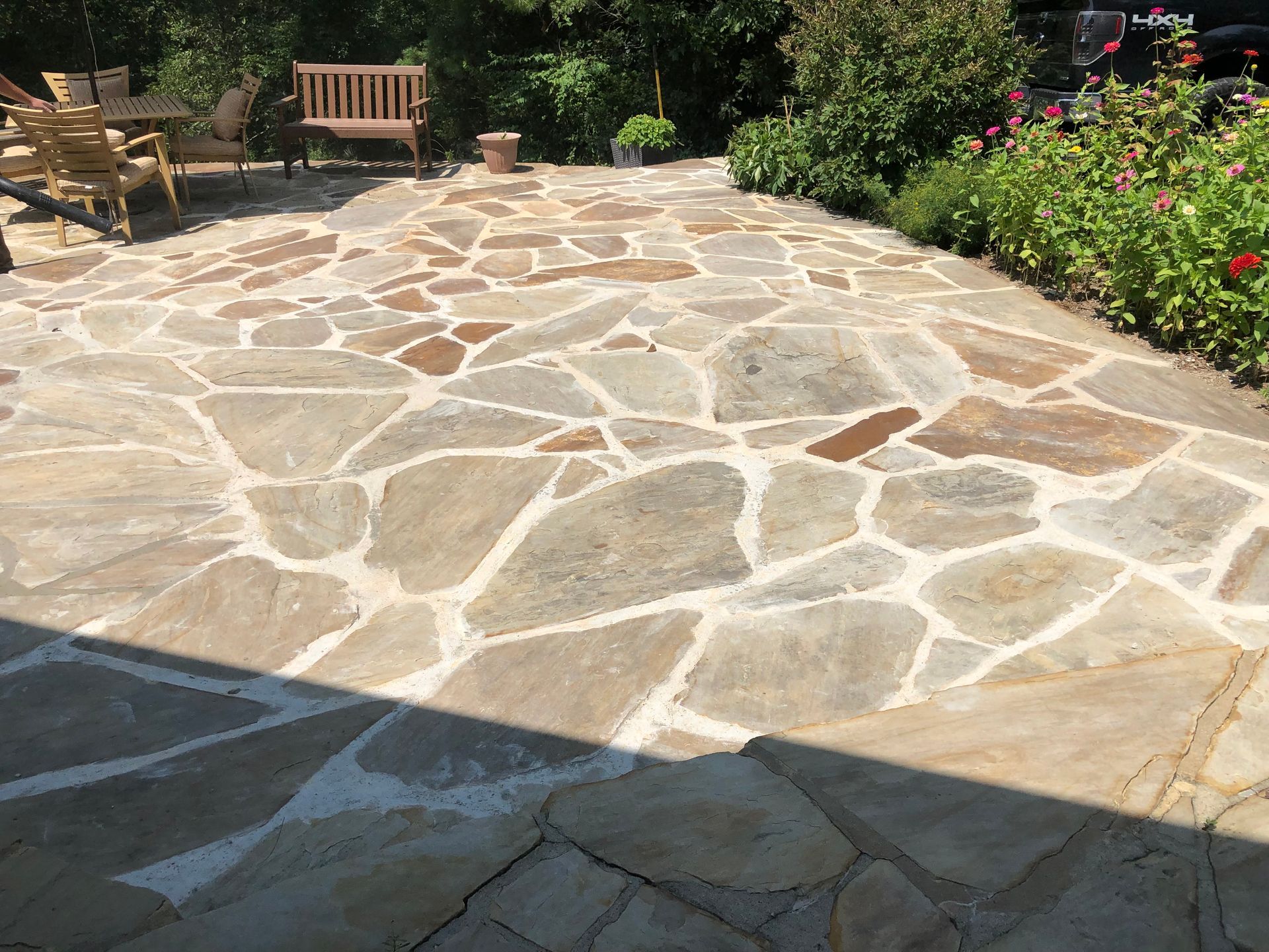 Stone patio with irregular flagstones, bordered by flowers and shrubs, with benches in the background