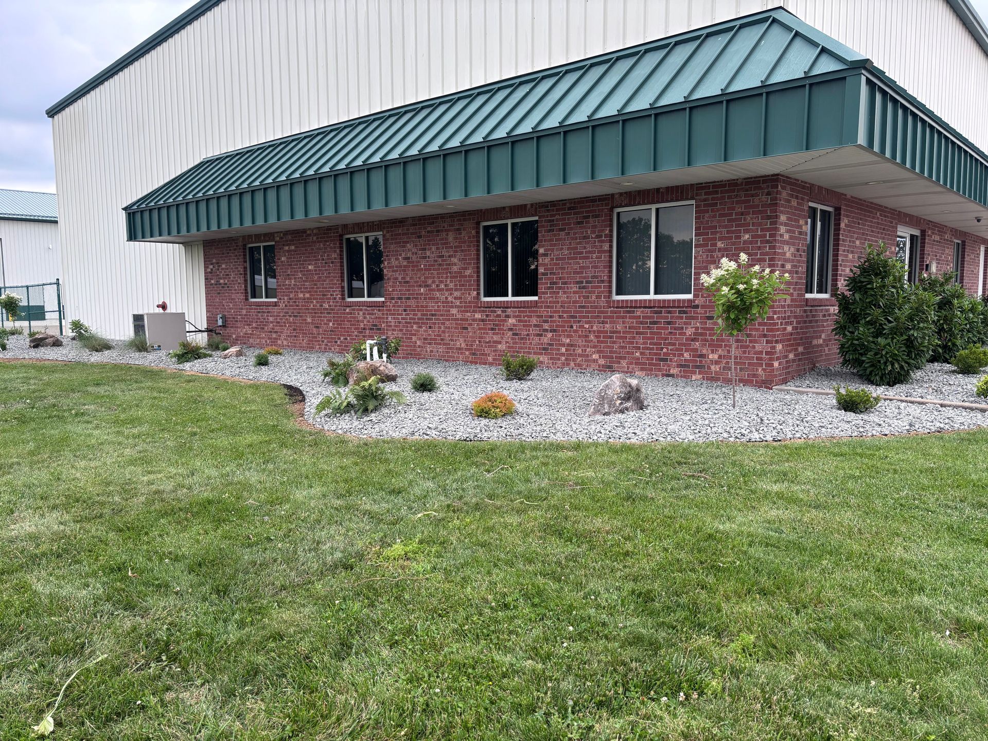 Brick commercial building with green metal roof and landscaped rock bed beside a grassy lawn