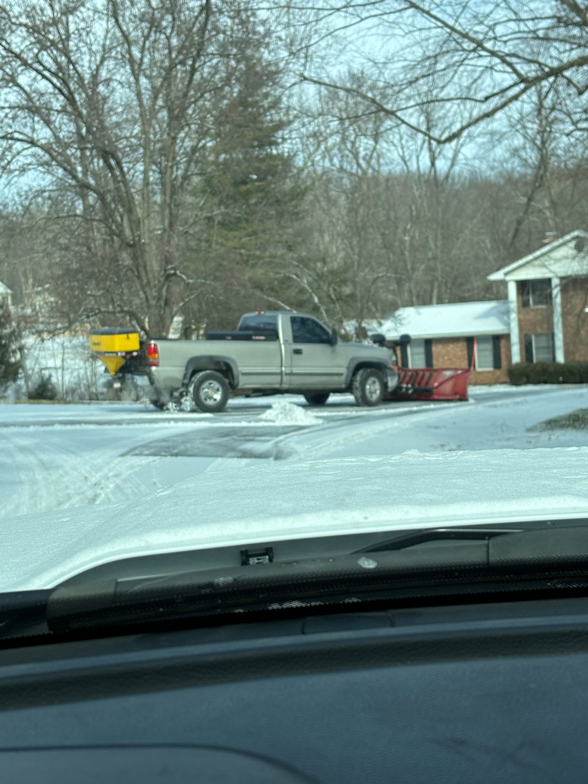 Pickup truck on a snowy driveway pulling a red snowplow, viewed from inside a car