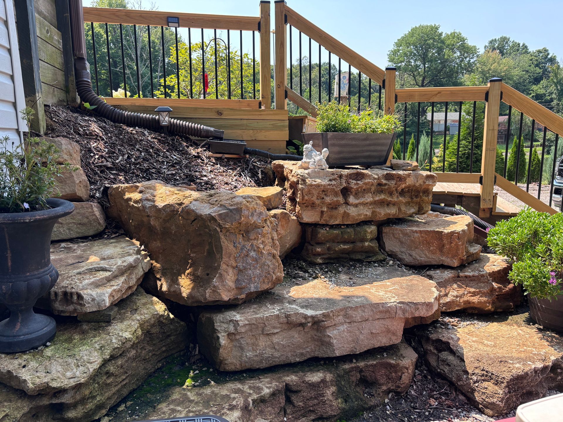 Stone retaining wall steps beside a wooden deck railing in a sunny garden
