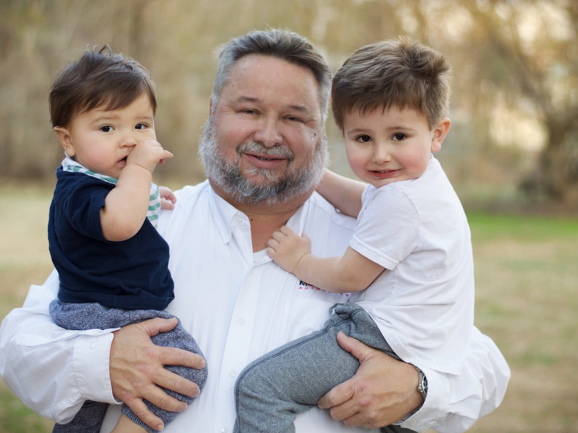Man holding two small children outdoors, all wearing casual clothes and smiling at the camera