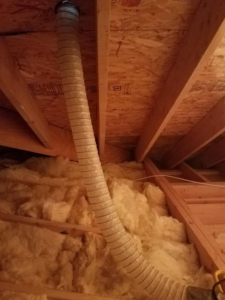 Inside a crawlspace, wooden beams and a dirt floor with white spray foam insulation on the back wall.