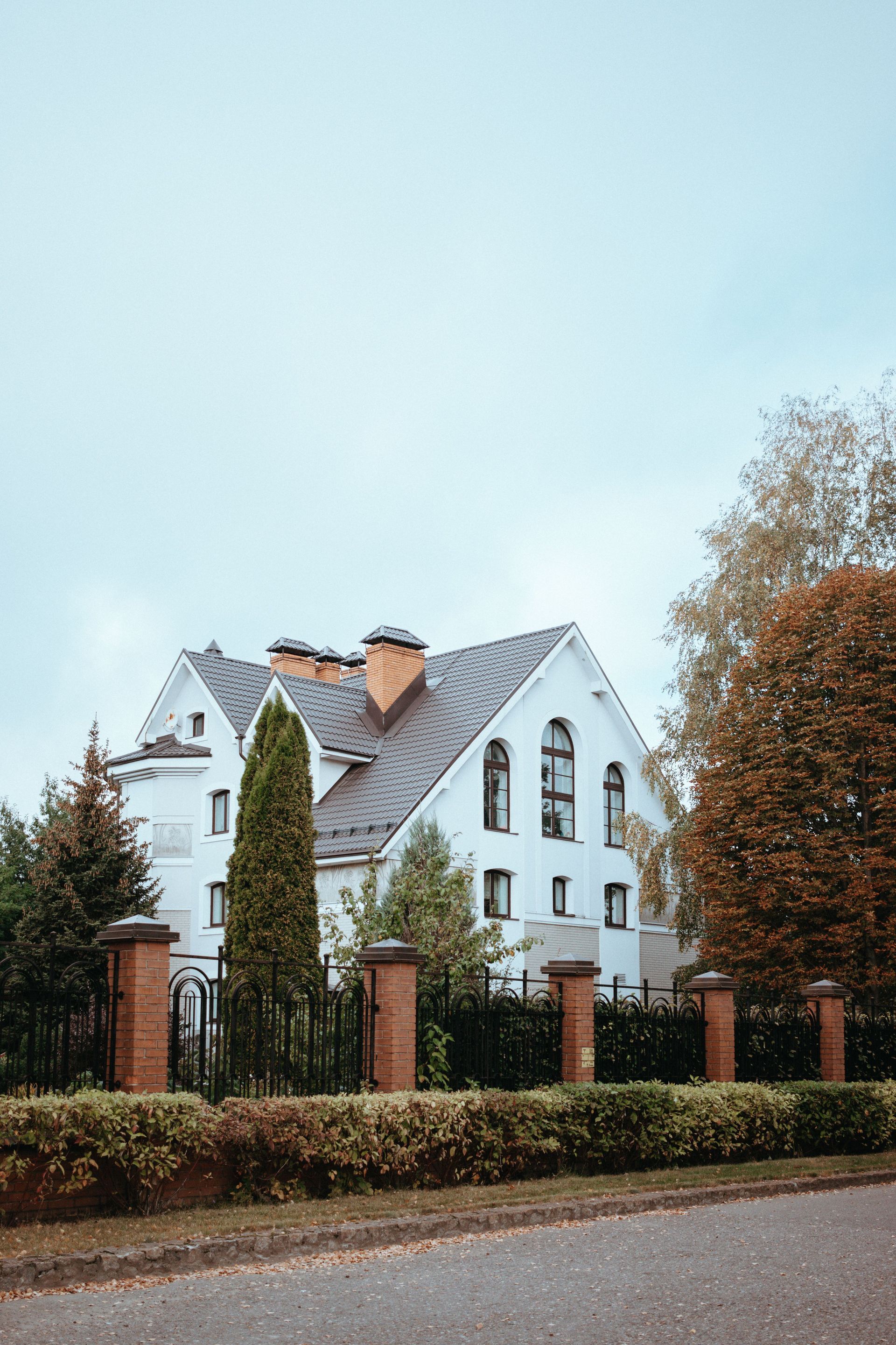 White house with a steep gray roof and brick fence, surrounded by trees under a pale sky