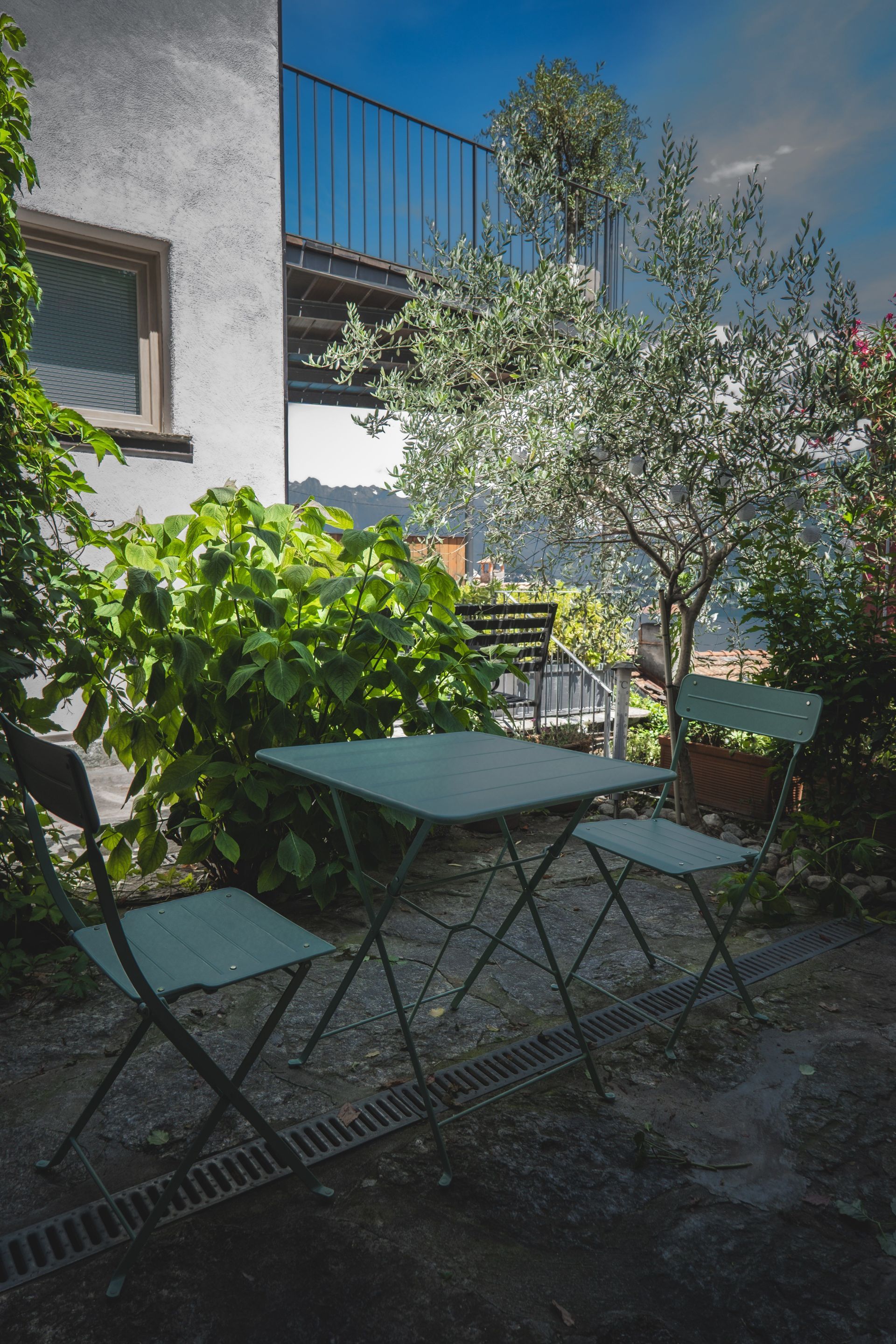 Small patio with a green table and chairs amid plants beside a white building