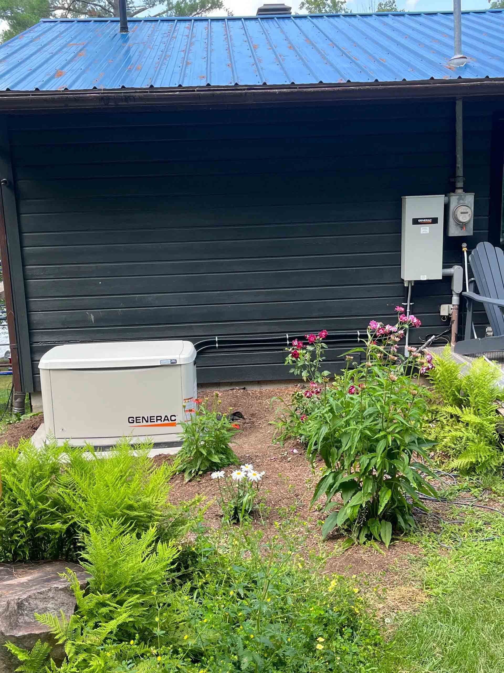 Generac generator installed in front of a garage with black doors and blue metal roof.