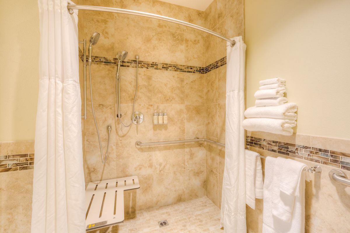 A walk-in shower with beige tile walls, a white shower curtain, a bench, and stacked white towels on a wall.