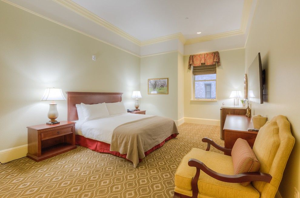 Hotel room with a double bed, two bedside tables with lamps, an armchair, and a desk. Beige walls and patterned carpet.