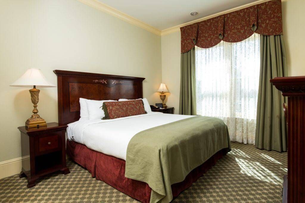 Hotel bedroom with a dark wooden bed and nightstands, a large window with curtains, and patterned carpet.
