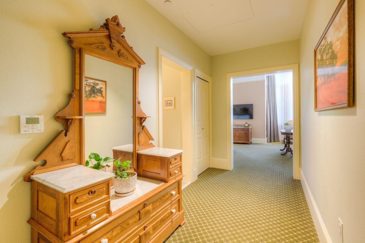 A hotel hallway with a decorative wooden dresser, ornate mirror, and doorway to a room with a TV.