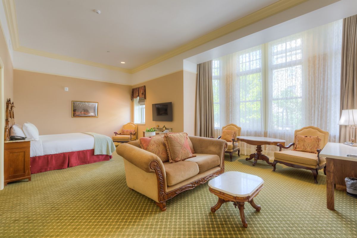 Spacious hotel room with a bed, sofa, two armchairs, a coffee table, and a large window. The room has antique furniture.