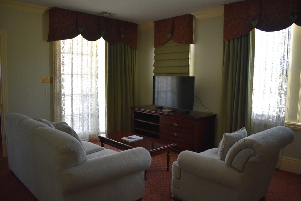 A hotel living room with a cream couch and armchair, TV, and windows with green drapes and patterned valances.