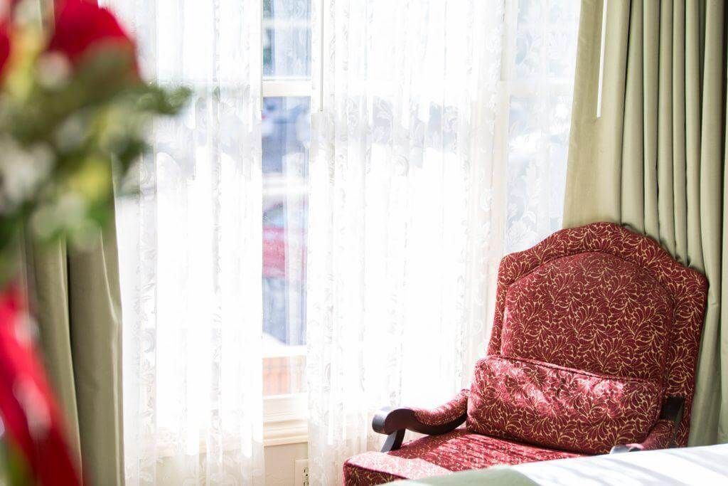 Red patterned armchair next to a window with sheer white curtains. Green curtains frame the right side.