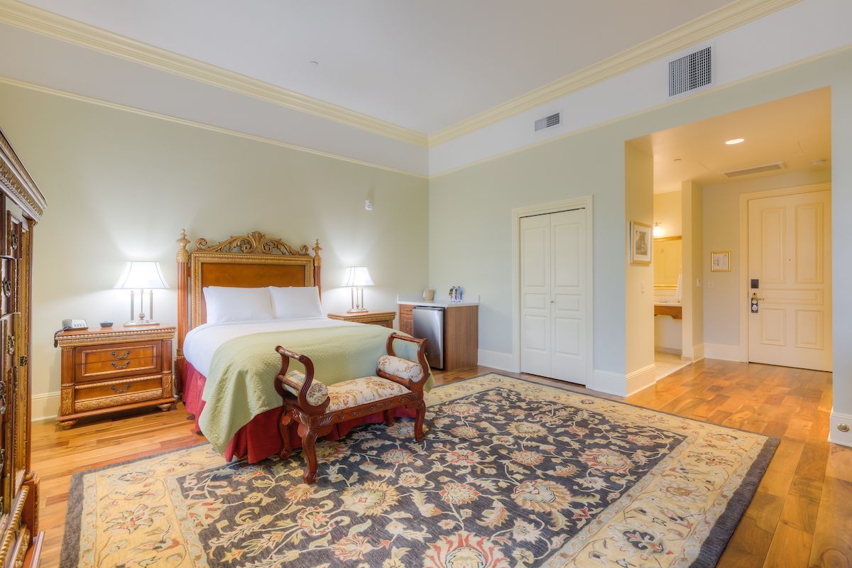A hotel bedroom with a bed, ornate furniture, rug, and an open doorway to a bathroom.