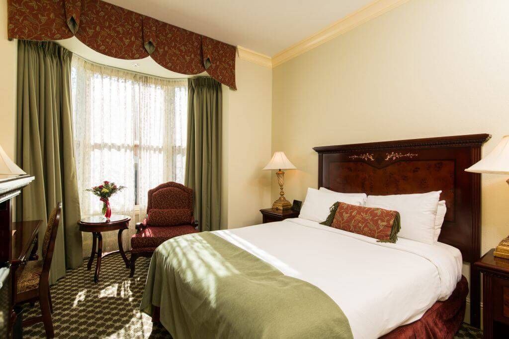 Hotel bedroom with a bed, a small table, a chair, and a bay window with curtains. Warm tones and traditional decor.