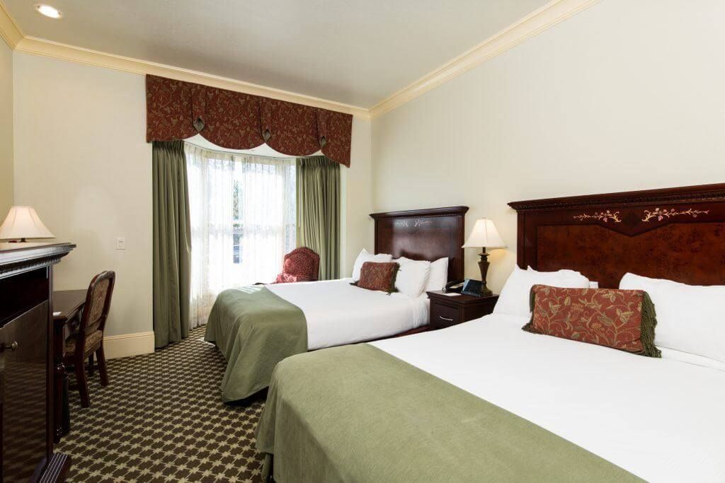 Two hotel beds with green bedspreads and decorative pillows in a room with a window and desk.