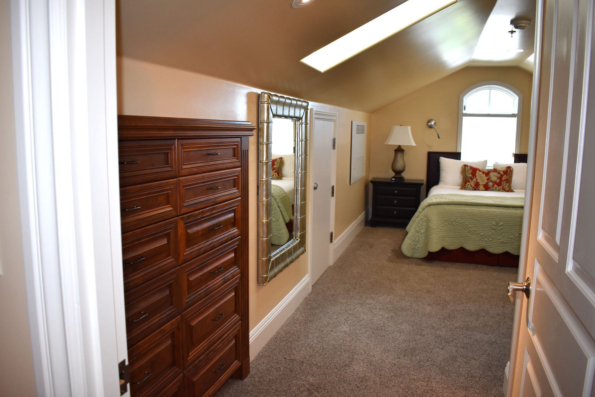 Bedroom with a dark wooden dresser, a long mirror, and a bed with a green quilt. The room has a sloped ceiling with a window.