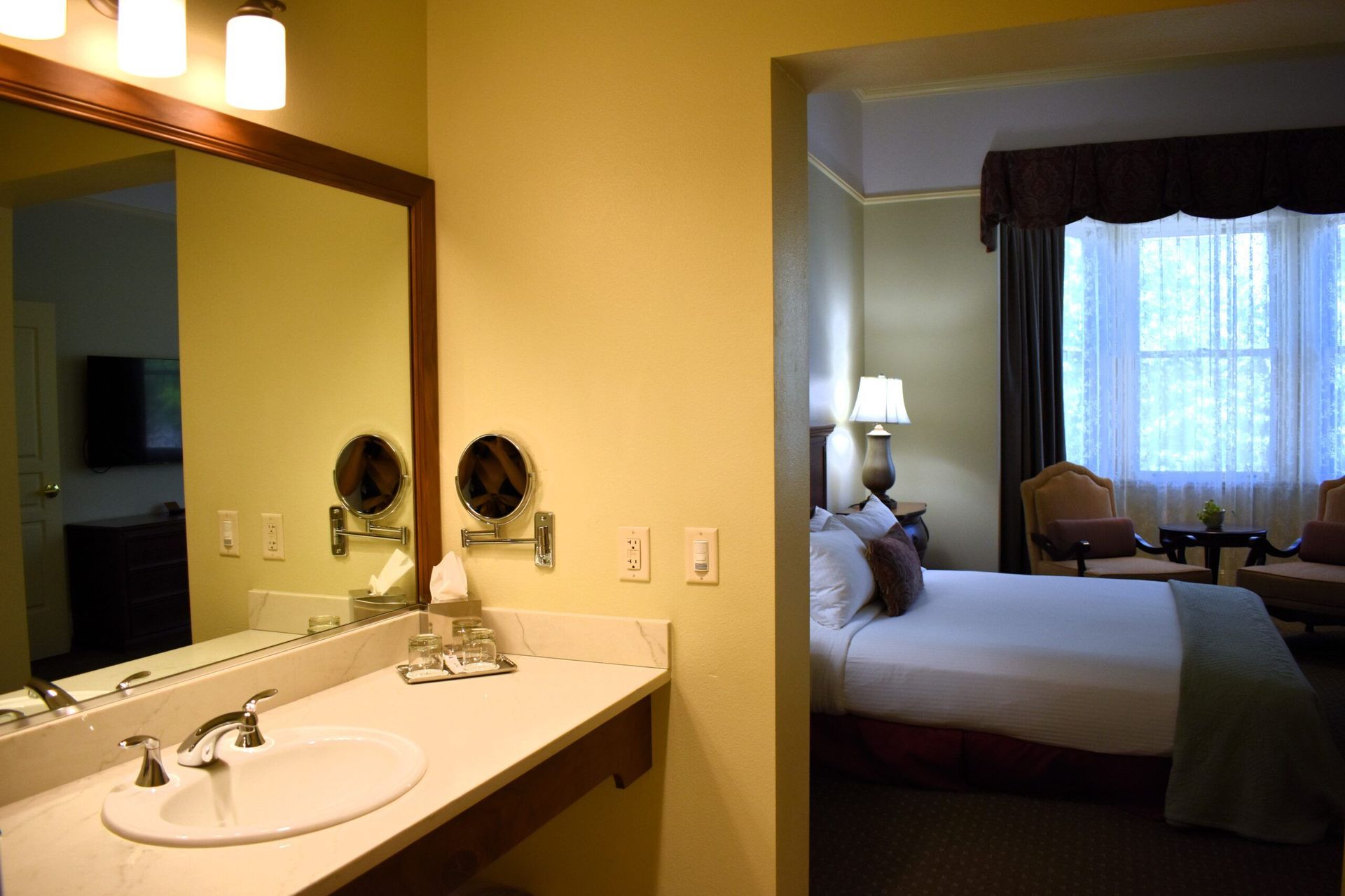 Bathroom with vanity and mirror, doorway to a hotel bedroom with a bed, chairs, and a window.